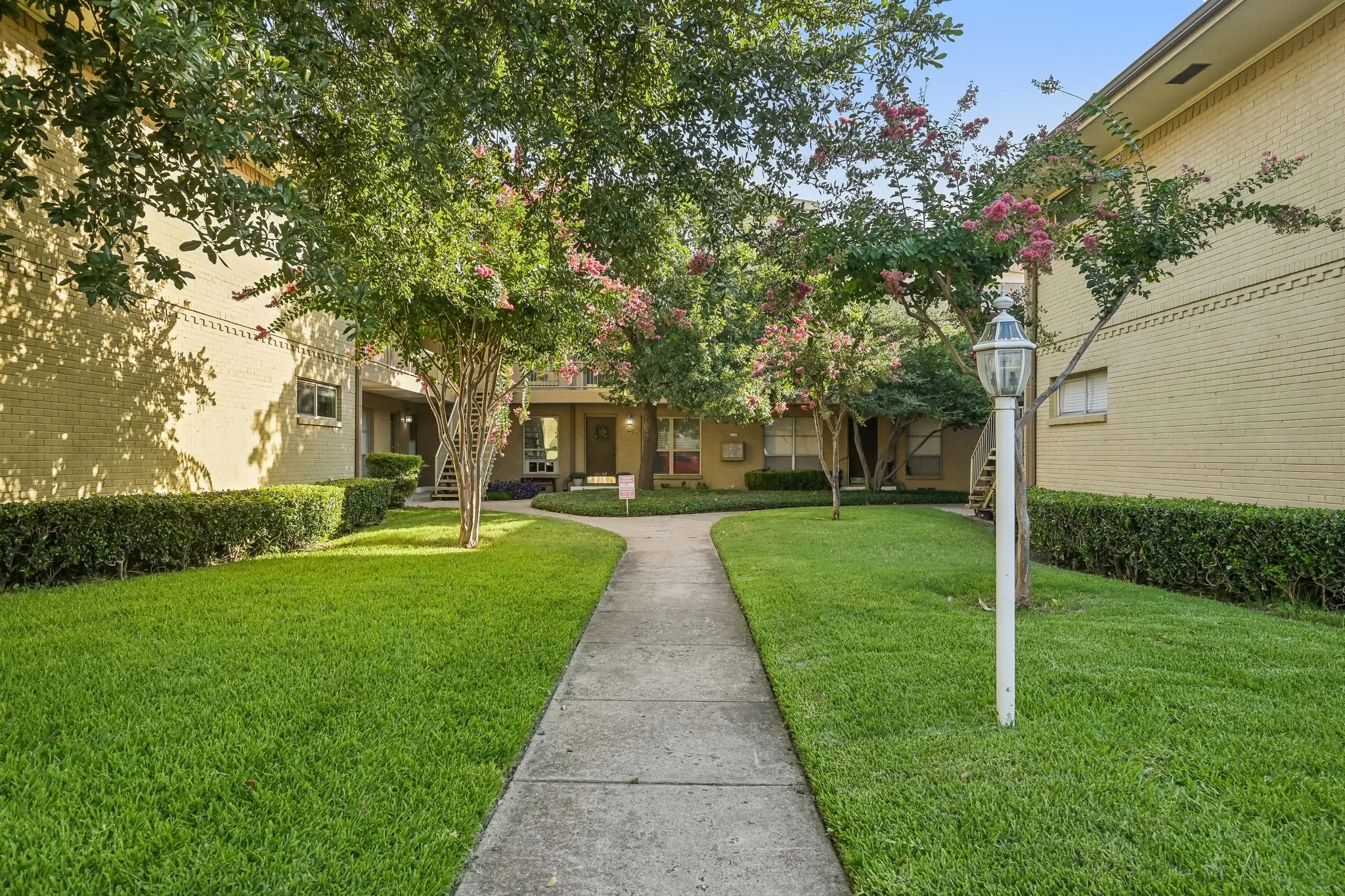 Well-maintained courtyard featuring a concrete pathway, manicured lawn, mature trees with pink blooms, and a classic white street lamp