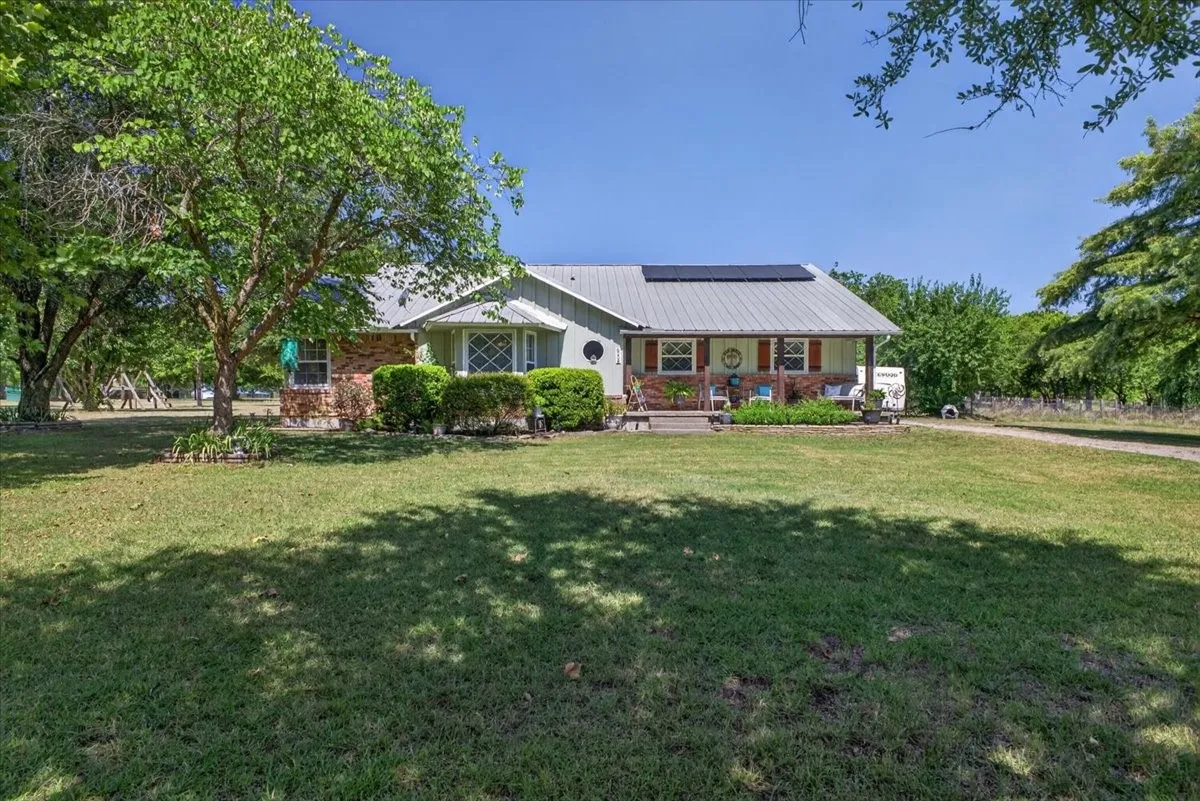 Front exterior with mature trees, metal roof, solar panels, and open front lawn