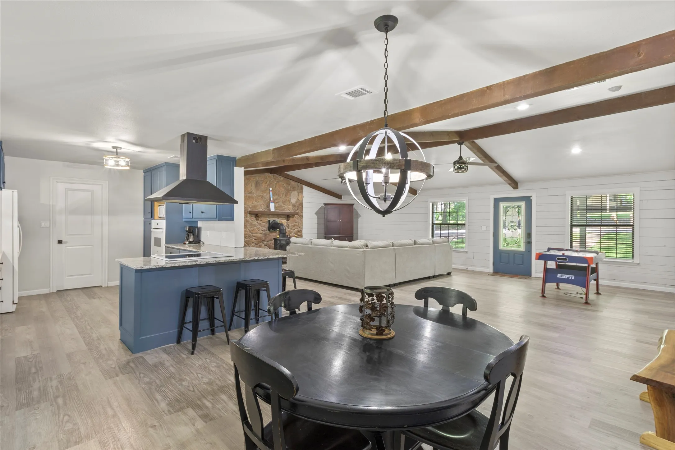 Open-concept living space with vaulted ceilings, exposed wood beams, wood-finish flooring, a kitchen island with stone countertop, and a stone accent wall