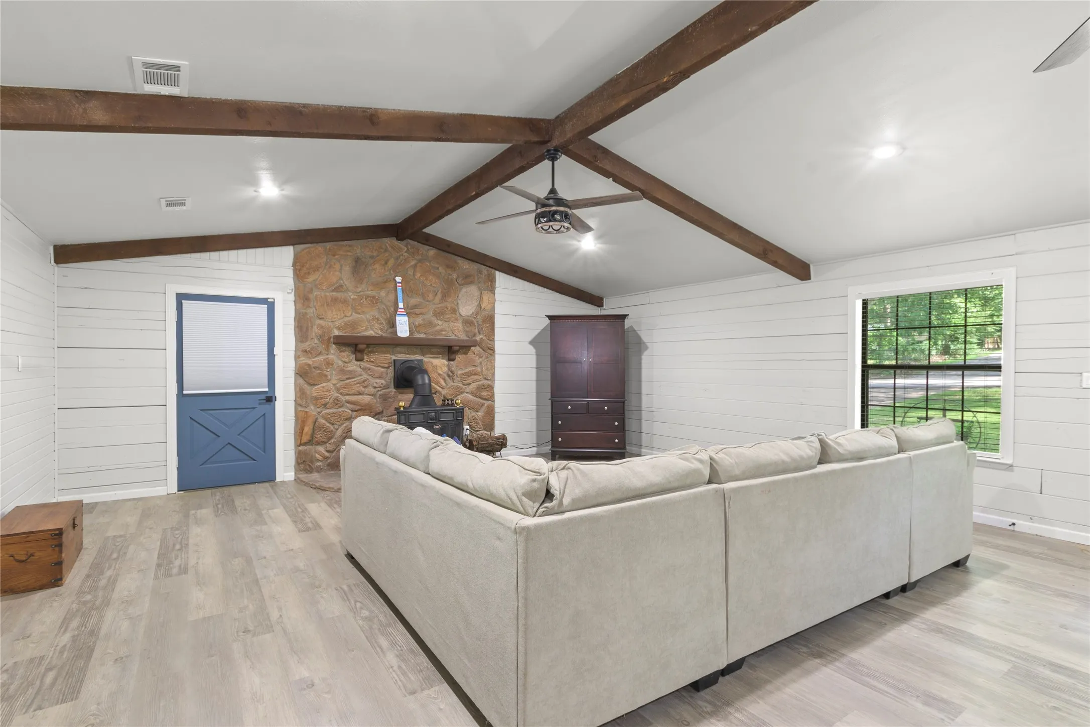 Vaulted ceiling with exposed wood beams, shiplap walls, stone-clad fireplace, wood-finish flooring, and a blue Dutch door