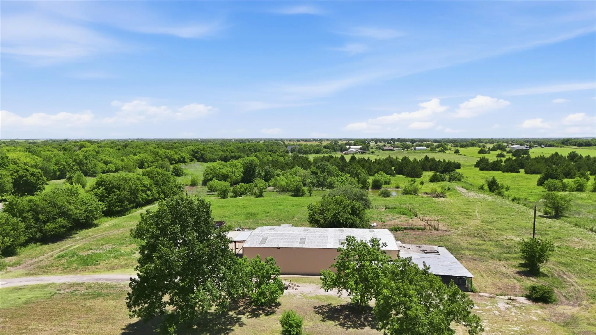 Expansive rural landscape featuring a single-story structure with a light-colored facade and a gray roof, surrounded by green fields and mature trees