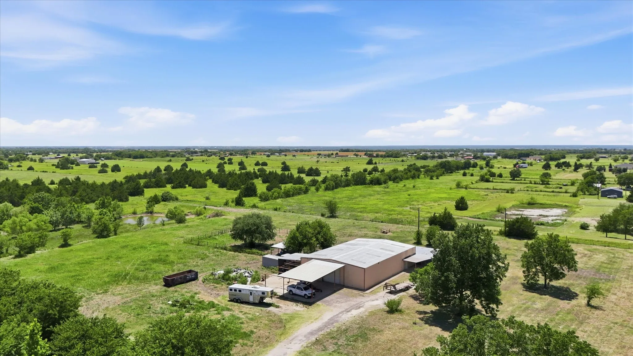 Expansive rural landscape featuring a large metal building with an attached carport, mature trees, and a distant pond