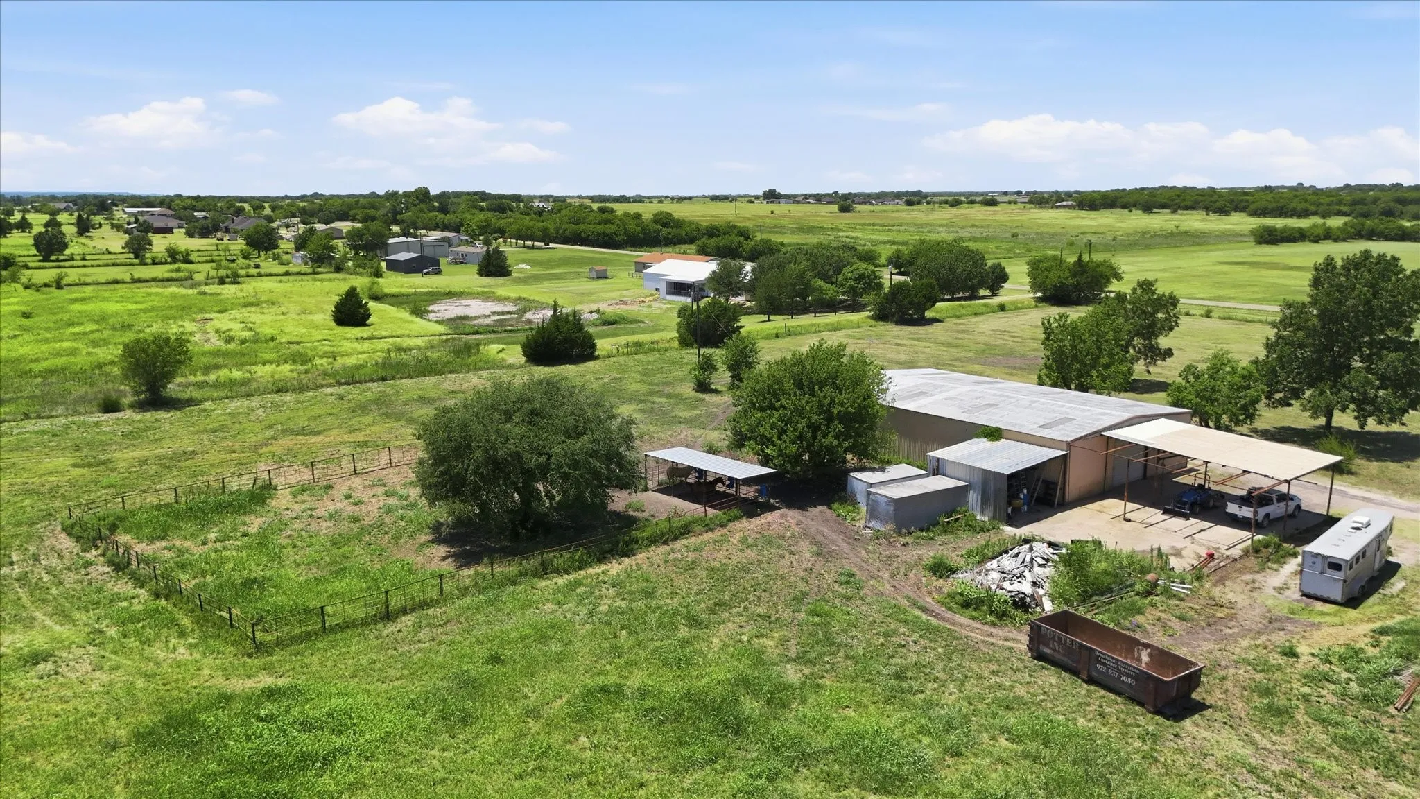 Expansive green acreage featuring a large metal building with an attached covered area, multiple outbuildings, and a fenced enclosure