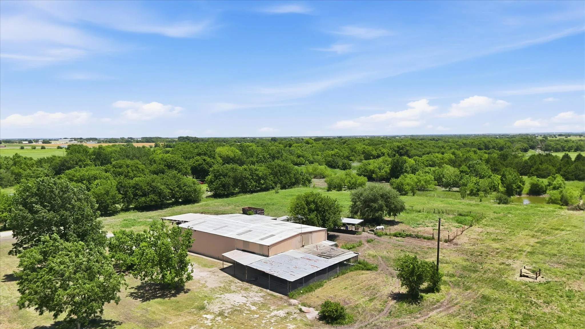 Expansive land featuring significant tree coverage, a large building with a metal roof, and an open-sided shed structure
