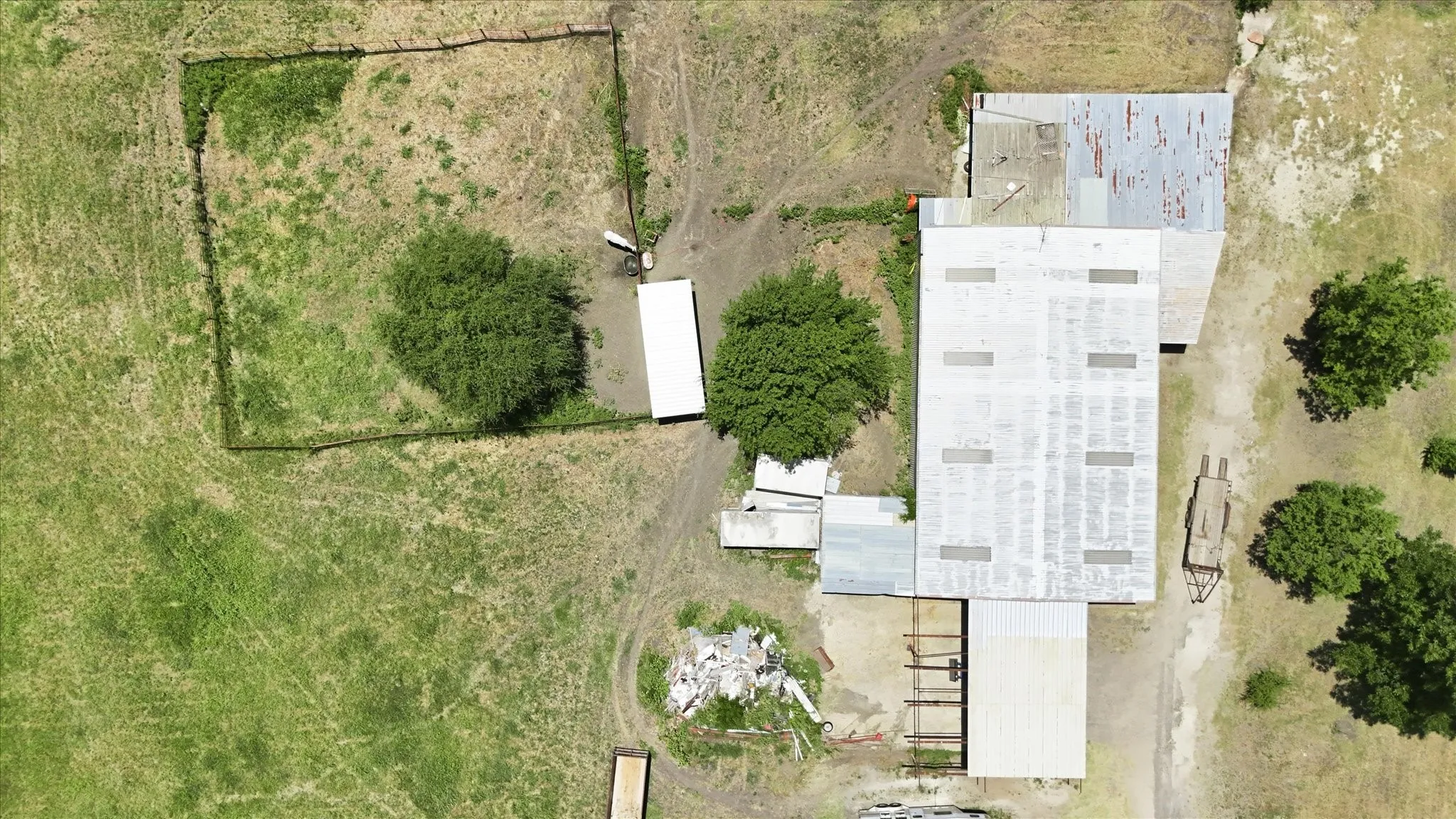 Extensive land parcel featuring a large primary structure with a light-colored roof, an additional rectangular building with a darker roof, and a fenced enclosure with a distinct green patch