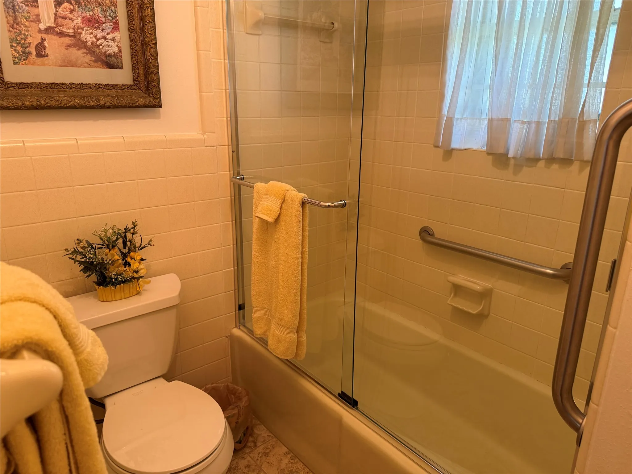 Bathtub with glass enclosure and grab bars, featuring tiled walls and a window with privacy curtain