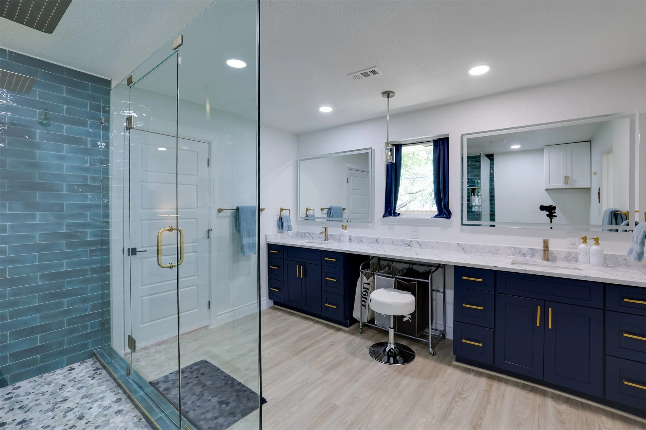 Spacious bathroom featuring a glass-enclosed shower with blue subway tile, a long double vanity with dark blue cabinetry, white marble-finish countertops, and dual wall-mounted mirrors