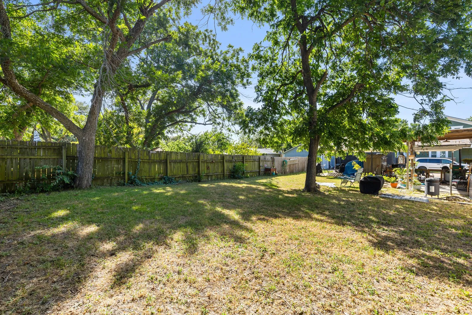 Spacious backyard with mature trees providing ample shade, bordered by a wooden privacy fence, and featuring a partially covered patio area