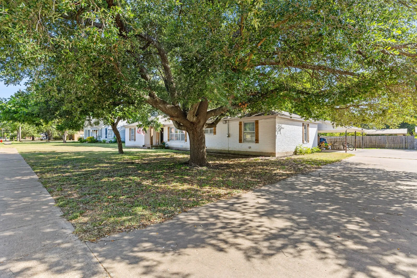 Mature shade trees canopy a white stucco home with brown shutters
