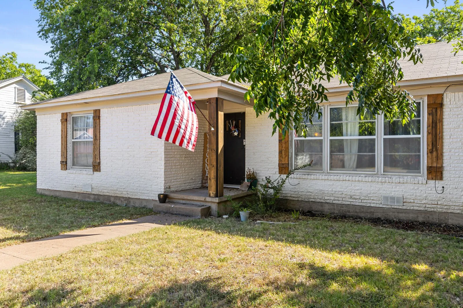White painted brick exterior with a covered front porch featuring a dark wood support post, a dark entry door, and multiple windows with wood-finish shutters