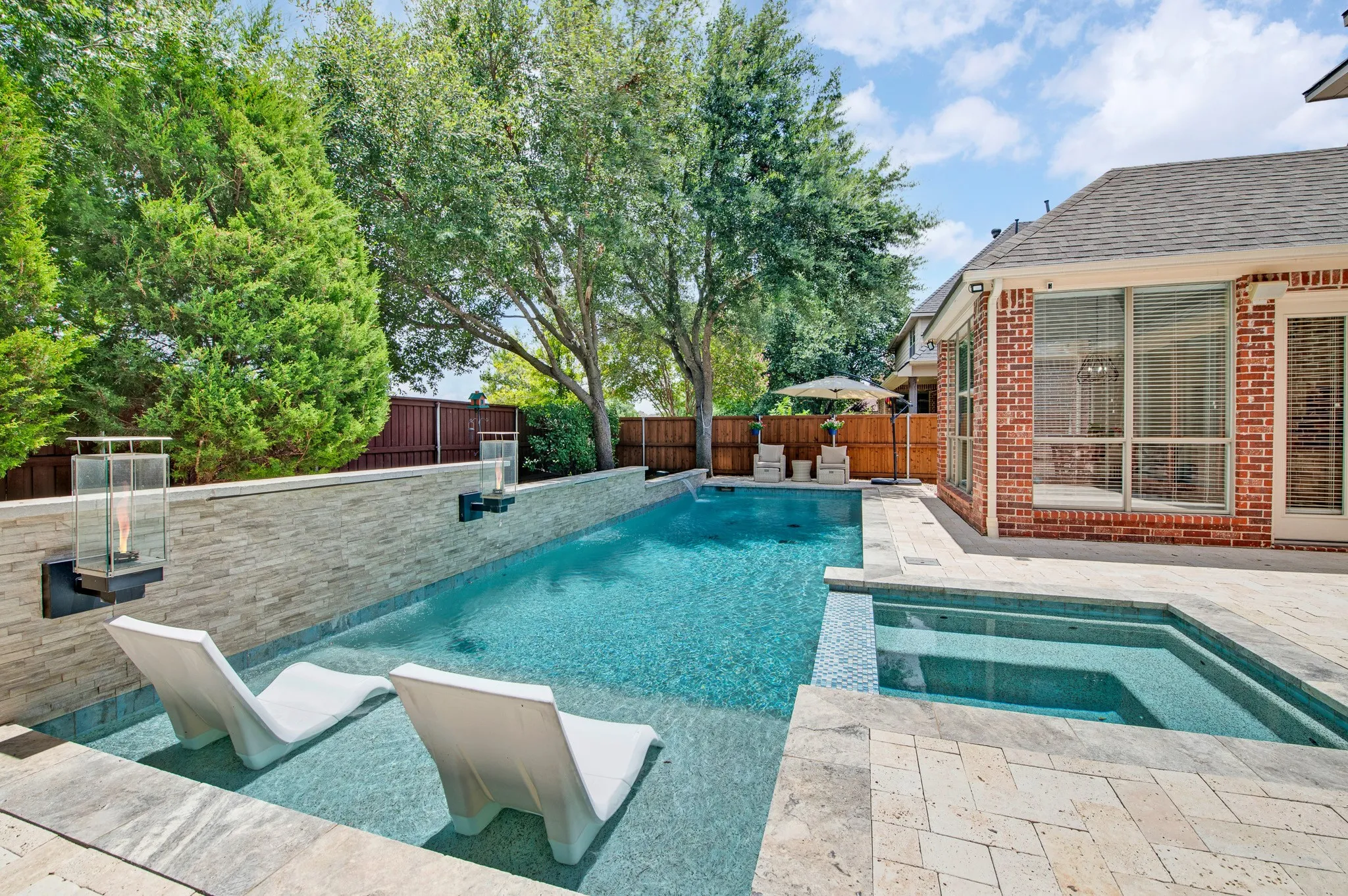 Backyard oasis featuring a rectangular swimming pool with integrated sun shelf, an elevated spa, and a stone-clad retaining wall with decorative sconces