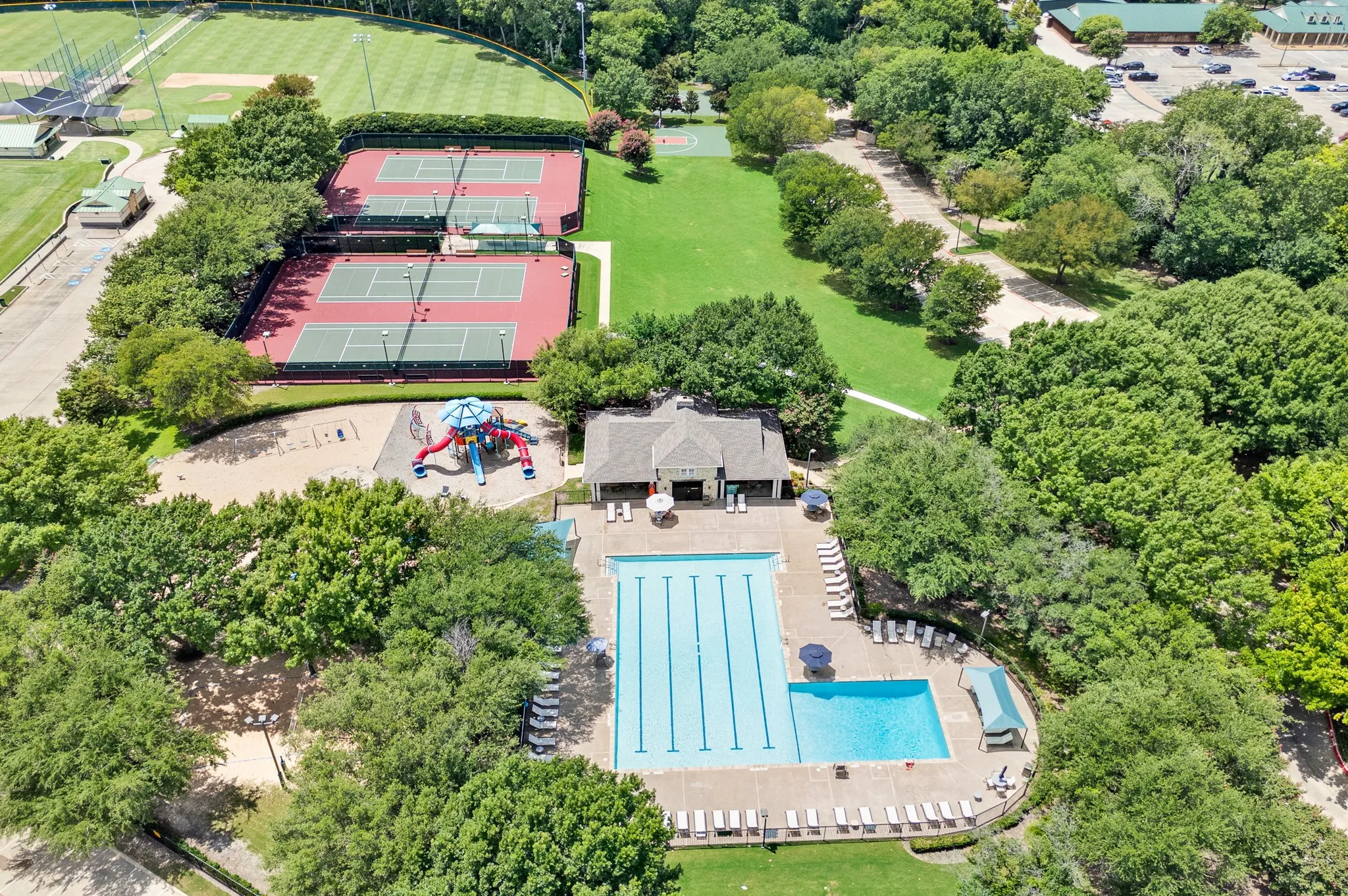 Community amenities featuring a multi-lane swimming pool with a separate shallow area, multiple tennis courts, a playground with sand, and expansive green spaces surrounded by mature trees