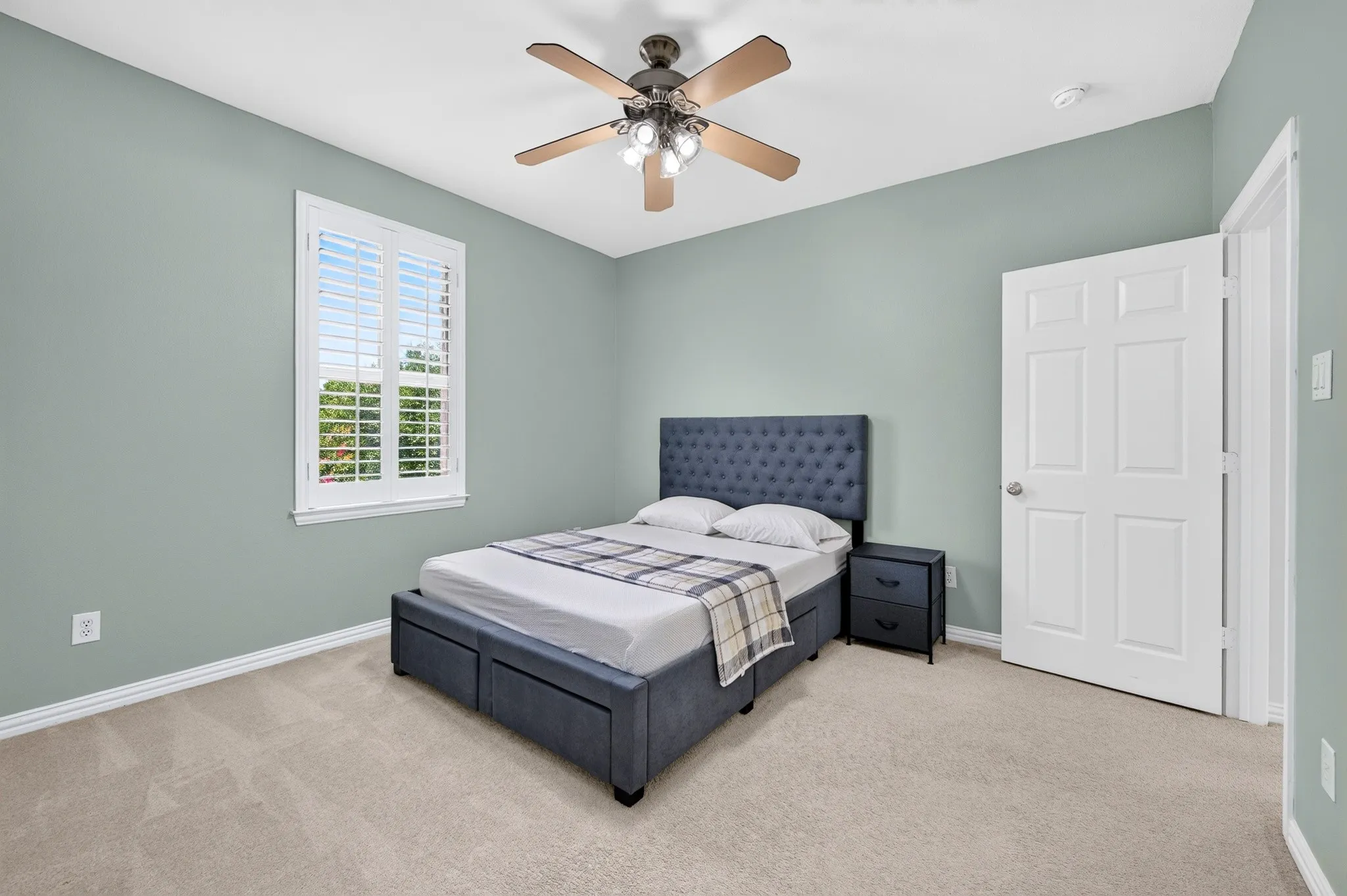 Carpeted bedroom featuring a window with white plantation shutters, a ceiling fan with light fixture, and light-toned walls