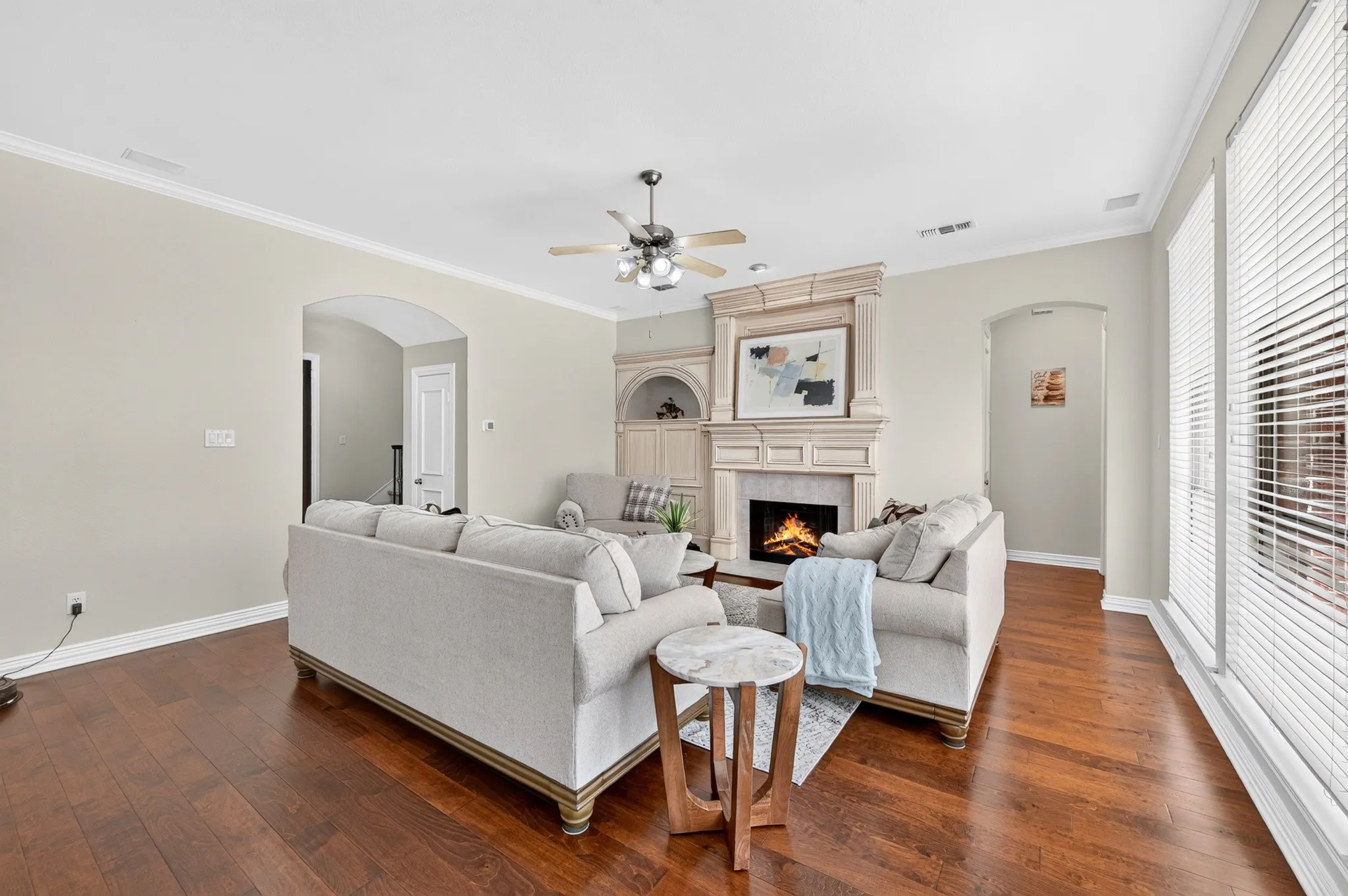 Living room featuring wood-finish flooring, a detailed fireplace with mantel, crown molding, arched doorways, and a ceiling fan
