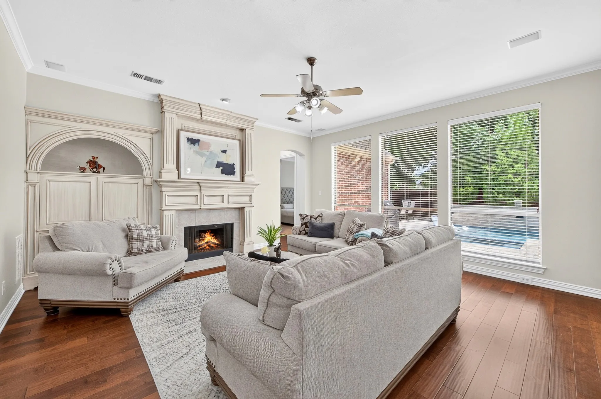 Living room featuring wood-finish flooring, a detailed built-in entertainment center, a stone-clad fireplace with mantel, and multiple large windows overlooking a pool