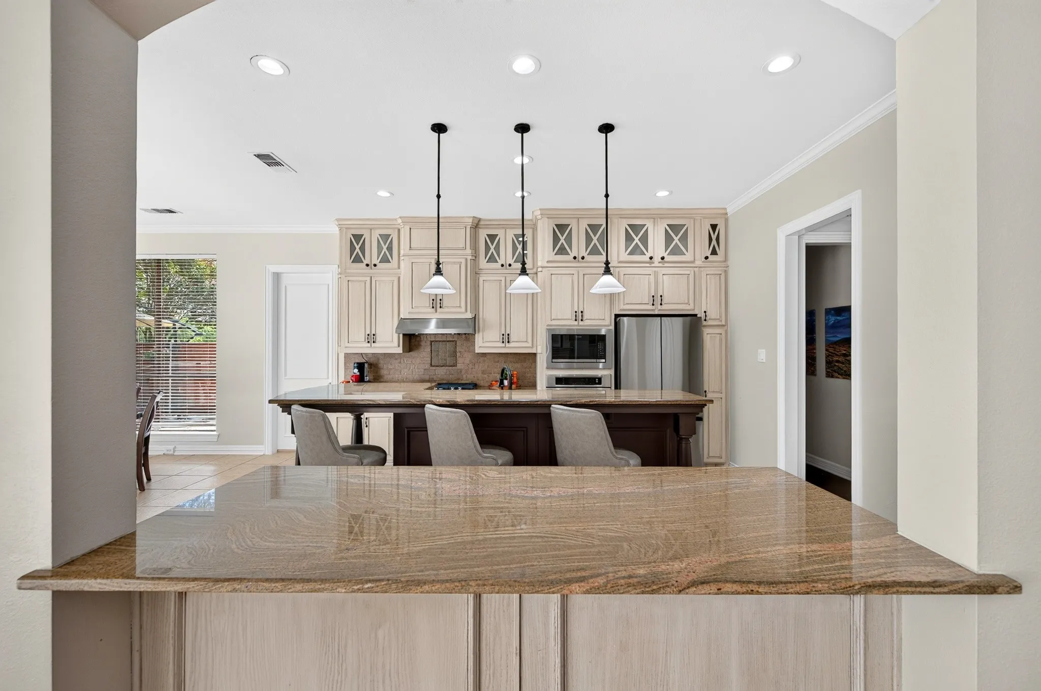 Kitchen featuring a large island with seating, pendant lighting, stainless steel appliances, light-toned cabinetry with glass-front accents, and recessed ceiling lights