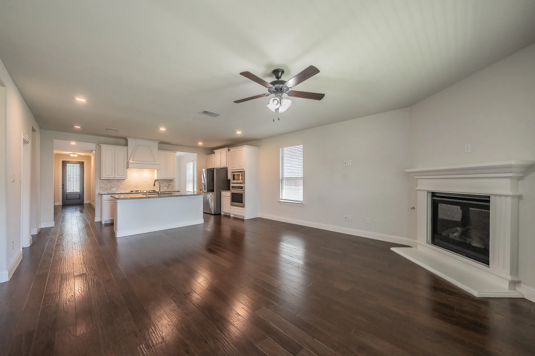 Expansive living area featuring rich wood-finish flooring, a white mantel fireplace, and a ceiling fan