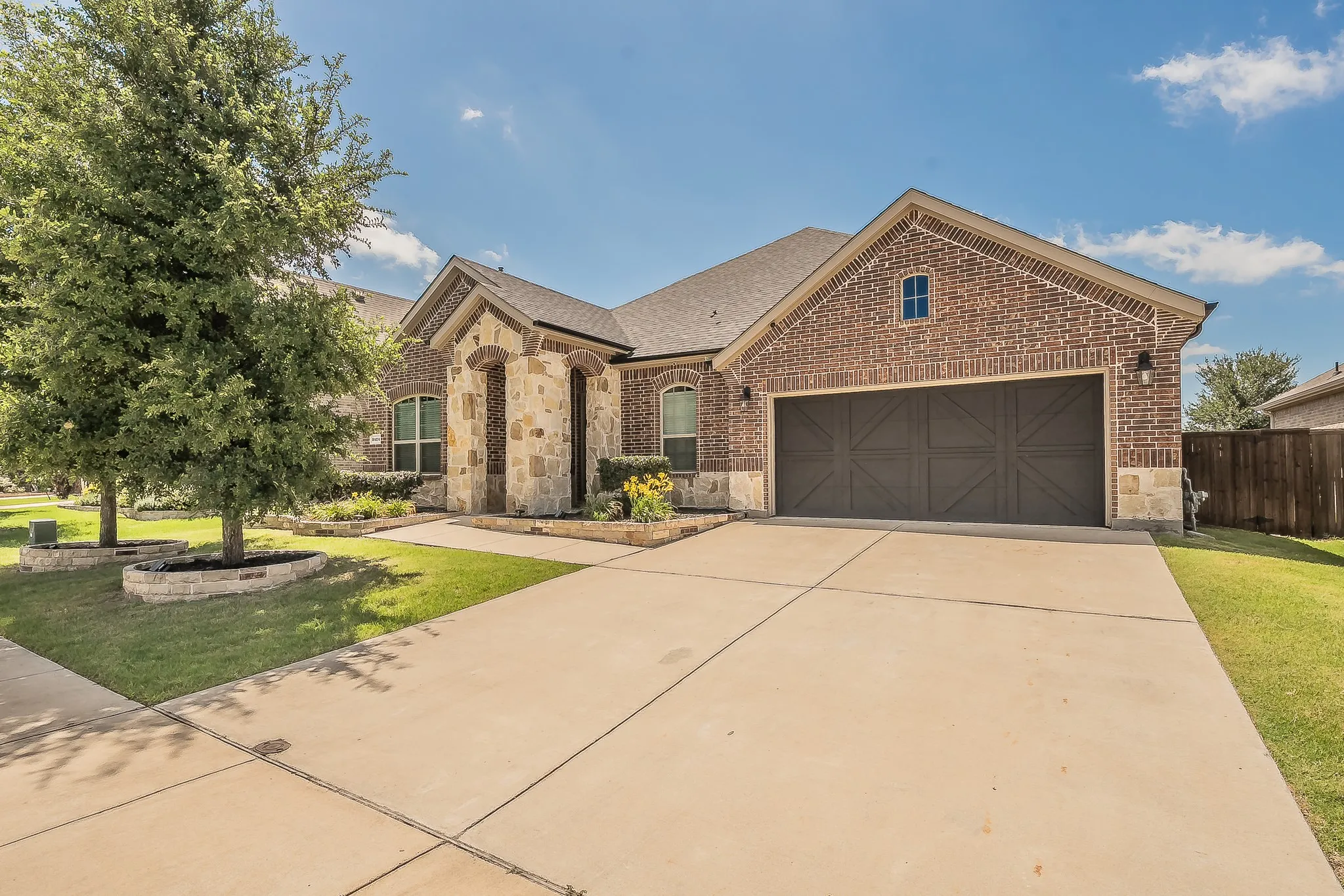 Brick and stone facade featuring a two-car garage with decorative doors, a concrete driveway, and established front landscaping with raised stone beds