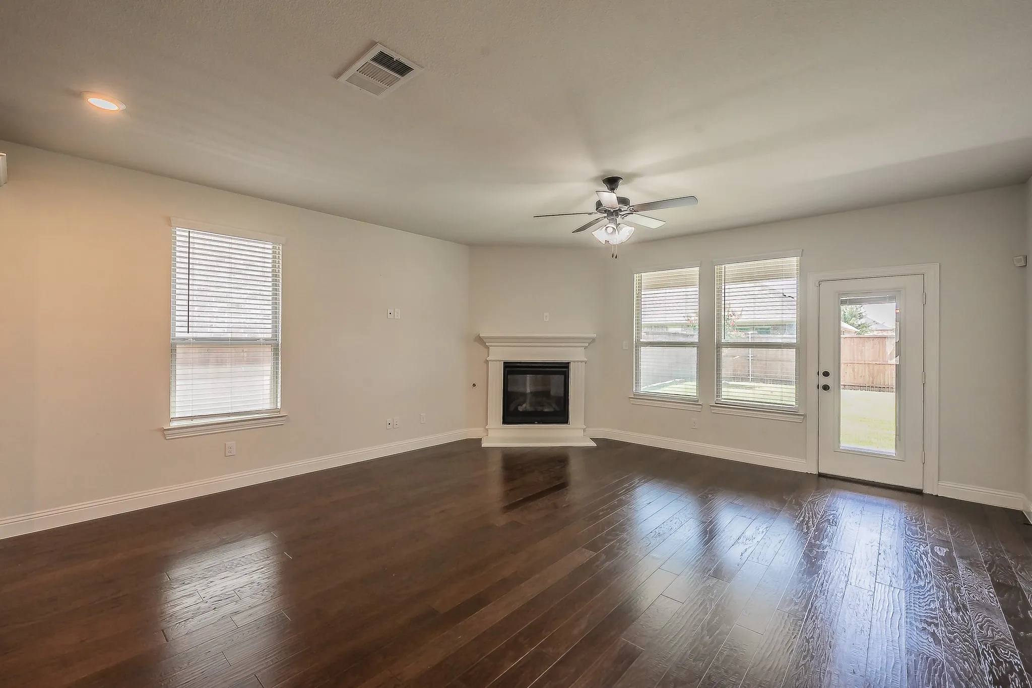 Spacious living area featuring rich wood-finish flooring, a corner fireplace with a white mantel, multiple windows with blinds, and a glass-paneled door opening to the exterior