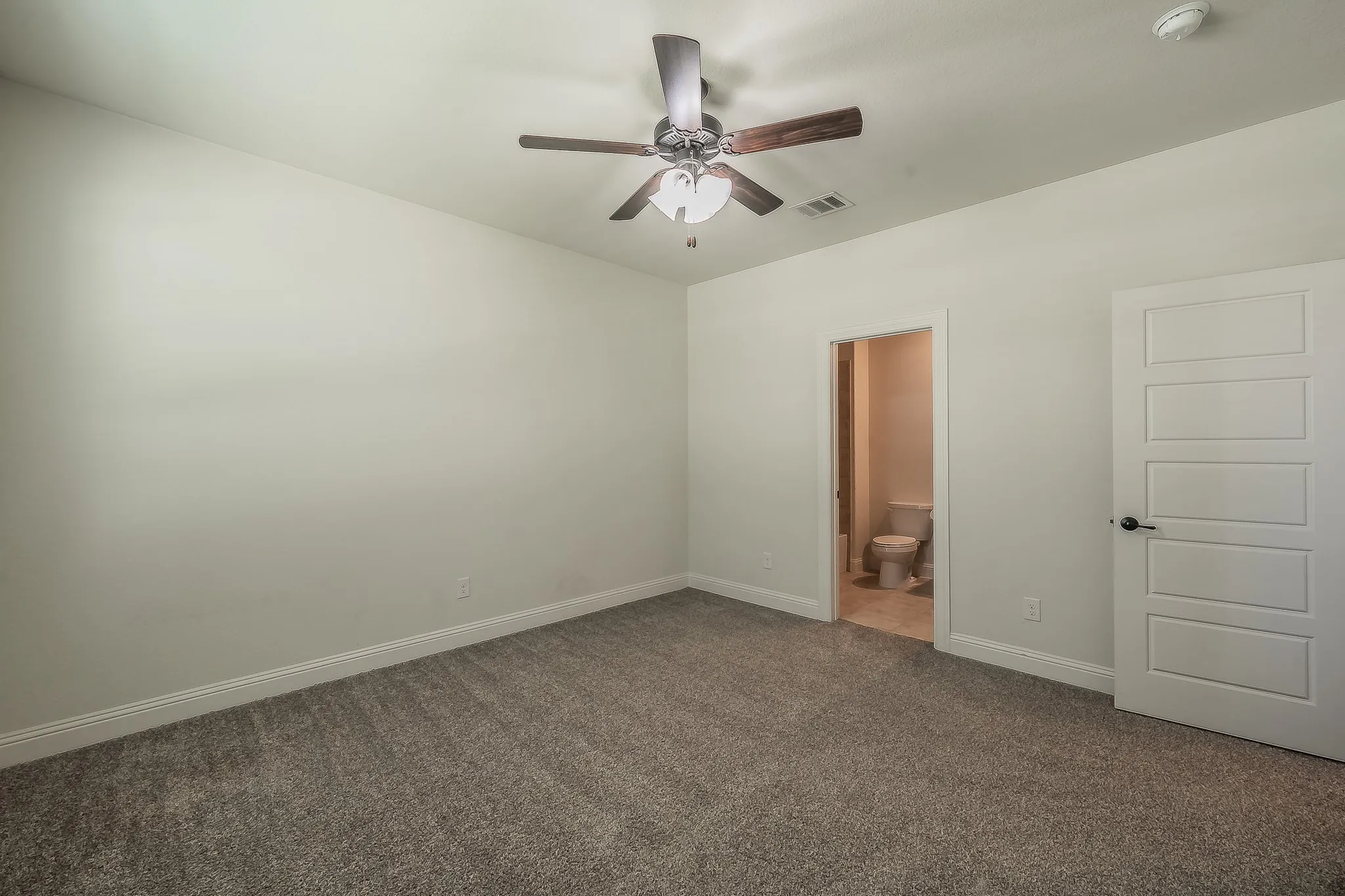 Carpeted interior room featuring a ceiling fan with light fixture, white five-panel interior door with matte black hardware, and an en-suite bathroom with toilet and tiled flooring