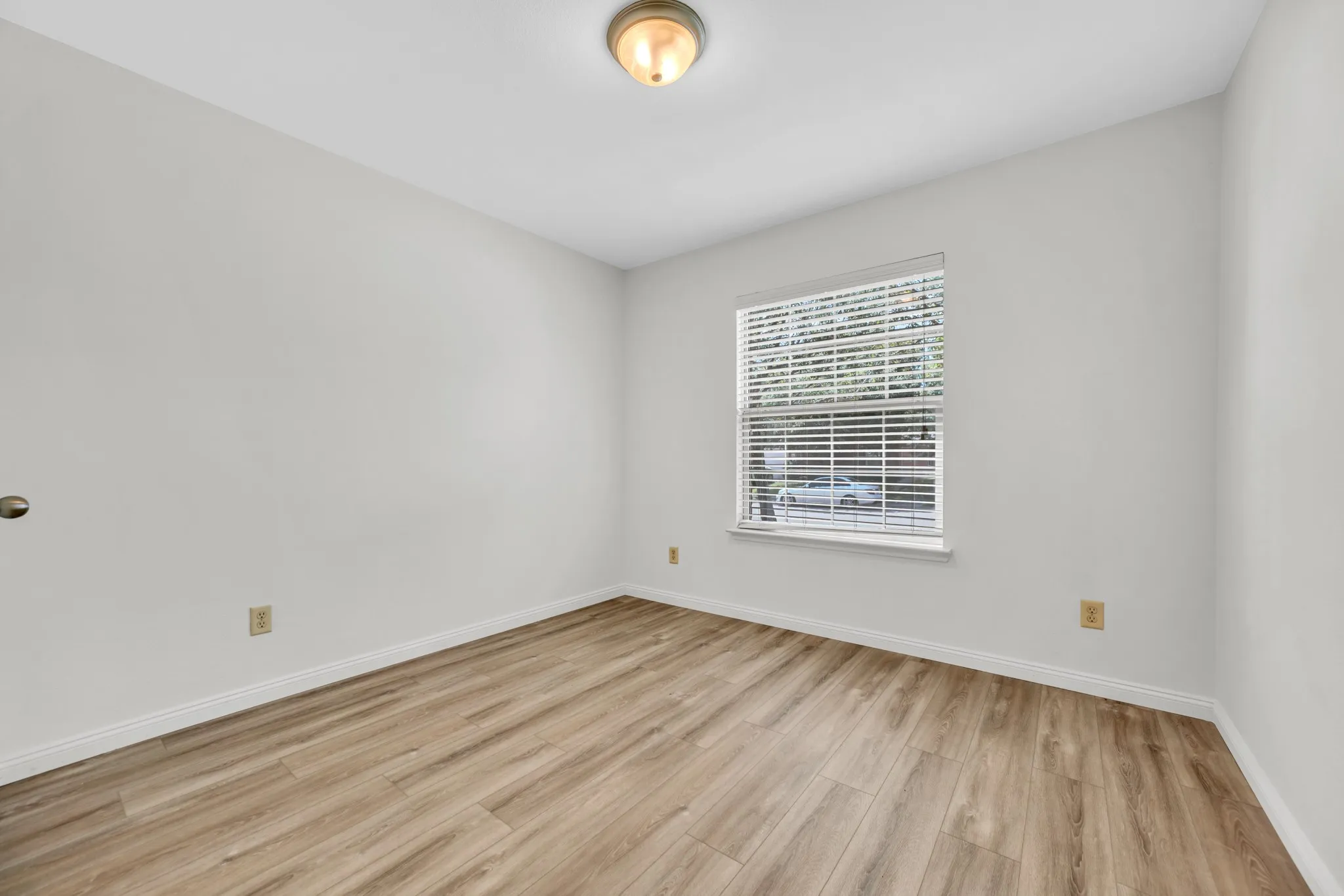 Room featuring wood-finish flooring, light-toned walls, a single window with blinds, and an overhead flush mount light fixture