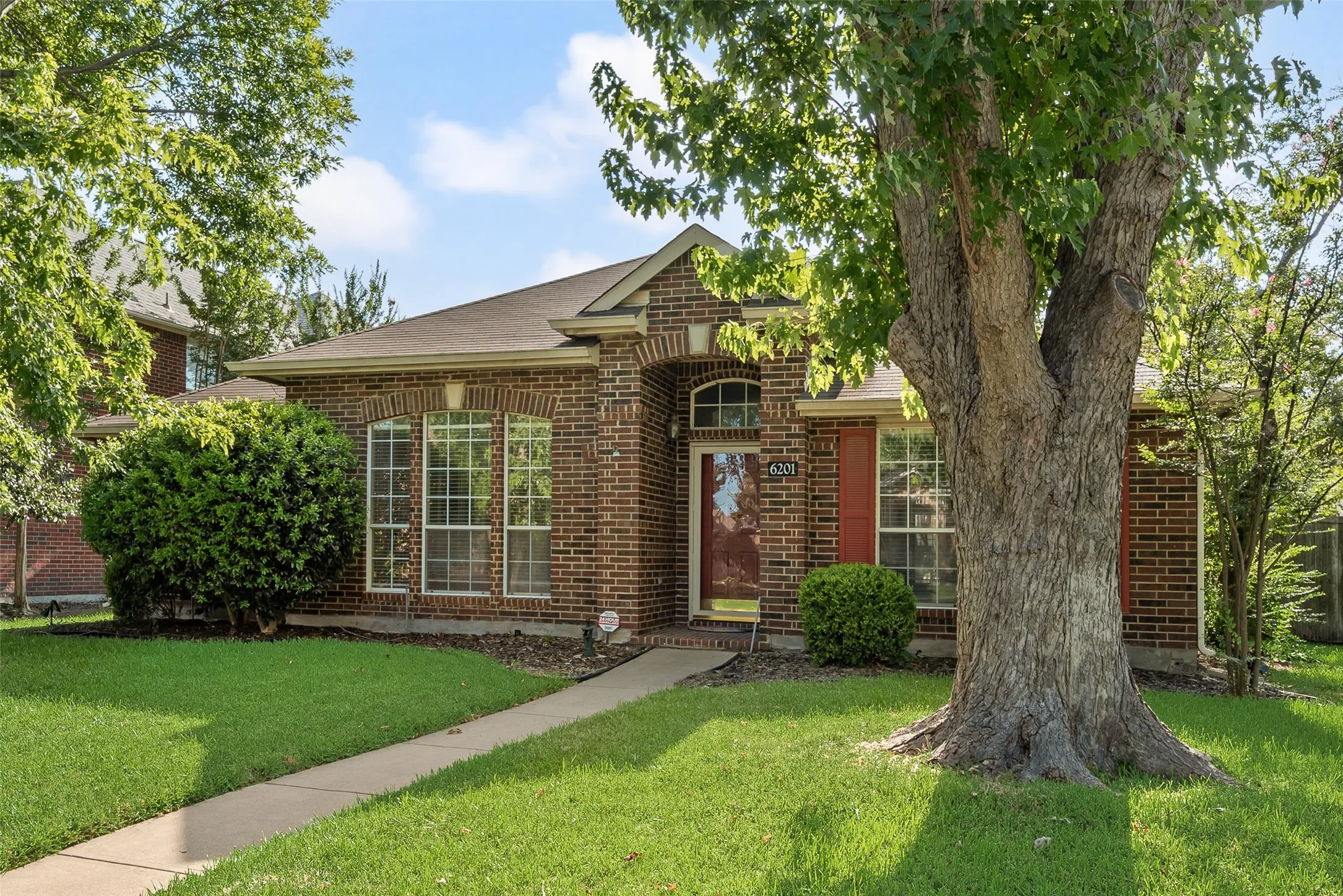 Brick exterior featuring a gabled roofline, a concrete pathway, mature trees, and landscaping