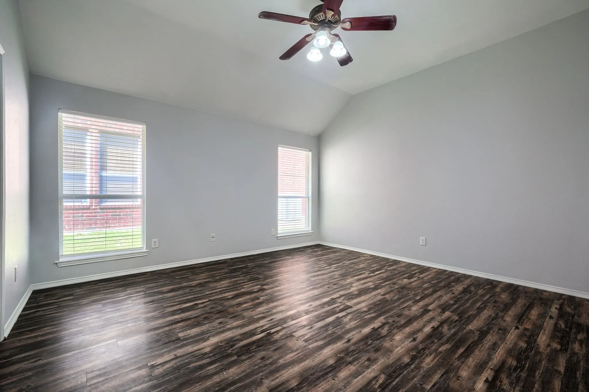 Room featuring vaulted ceilings, wood-finish flooring, two window openings with blinds, a ceiling fan with integrated lighting, and neutral wall paint