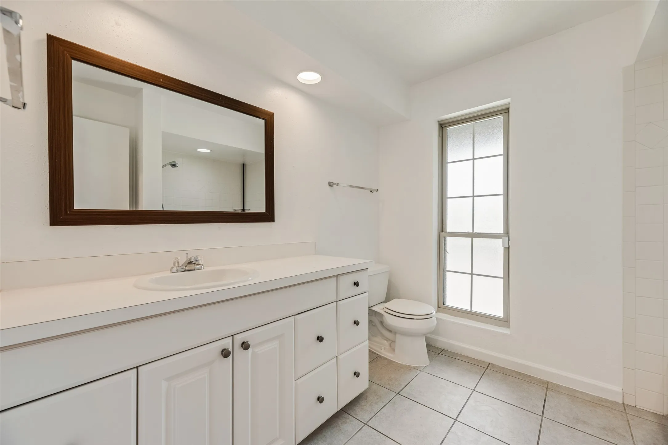 Bathroom featuring a white vanity with integrated sink, a rectangular framed mirror, tile flooring, a window with frosted panes, and a commode