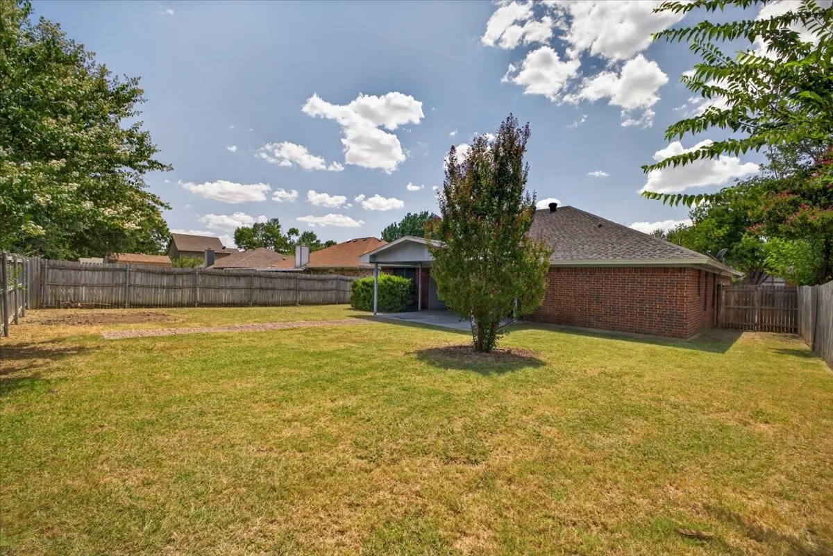 Expansive backyard featuring a mature tree, brick exterior, covered patio, and wood privacy fencing