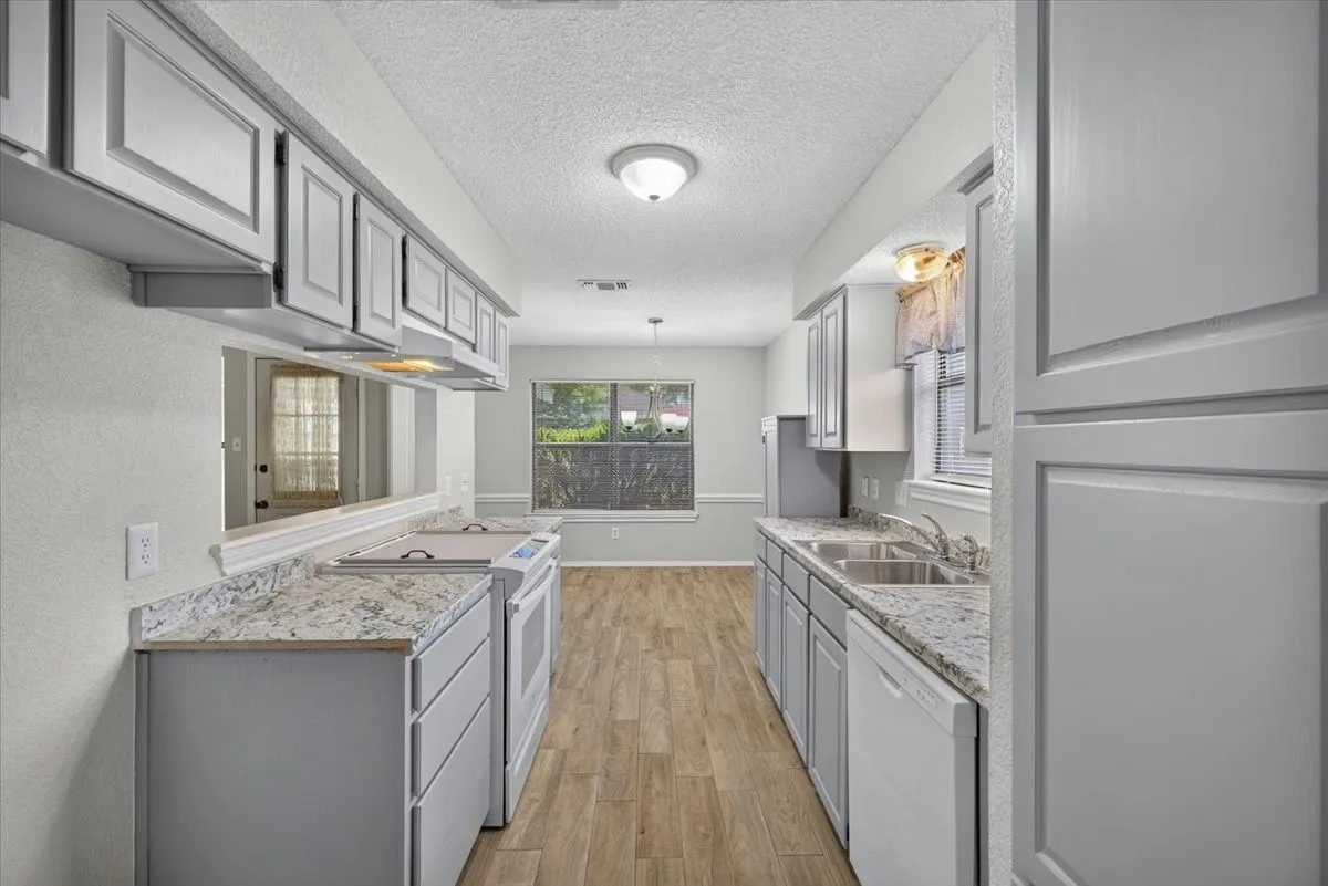 Galley kitchen featuring light gray cabinetry, light-toned countertops, wood-finish flooring, a double basin sink, and a built-in dishwasher