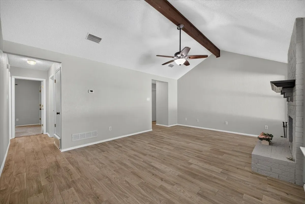 Living area featuring wood-finish flooring, a vaulted ceiling with exposed beams, a ceiling fan, and a brick fireplace with a mantel