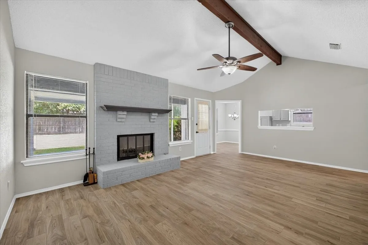 Spacious living area featuring a vaulted ceiling with exposed wood beam, wood-finish flooring, and a gray-painted brick fireplace with a wood mantle