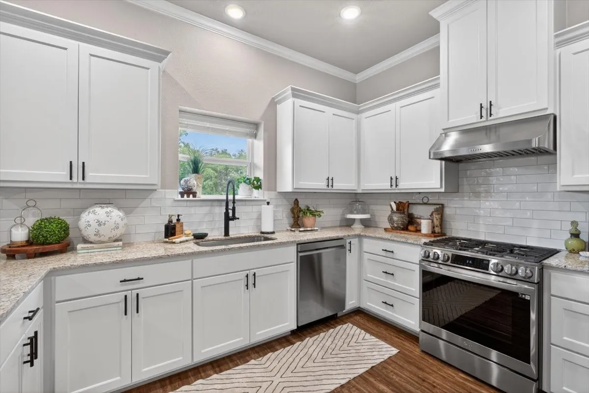 Modern kitchen featuring white shaker-style cabinetry, light-toned countertops, a subway tile backsplash, and stainless steel appliances including a gas range, dishwasher, and range hood