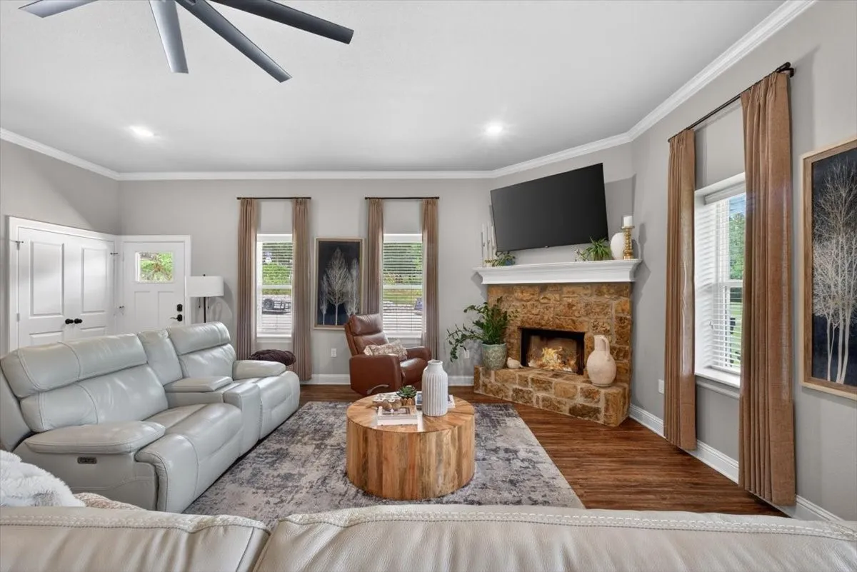 Living area featuring a stone-clad fireplace with a white mantle, wood-finish flooring, recessed lighting, crown molding, and a ceiling fan