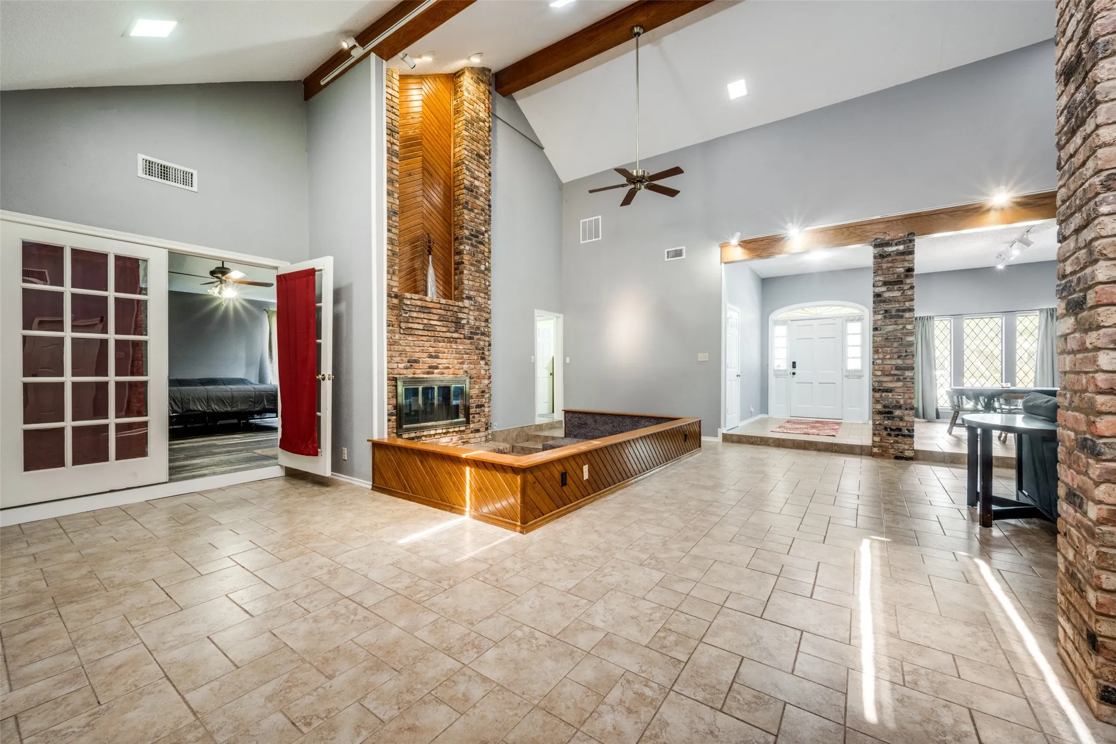 Vaulted ceilings with exposed wood beams, a brick fireplace with a wood-paneled surround, and tile flooring throughout