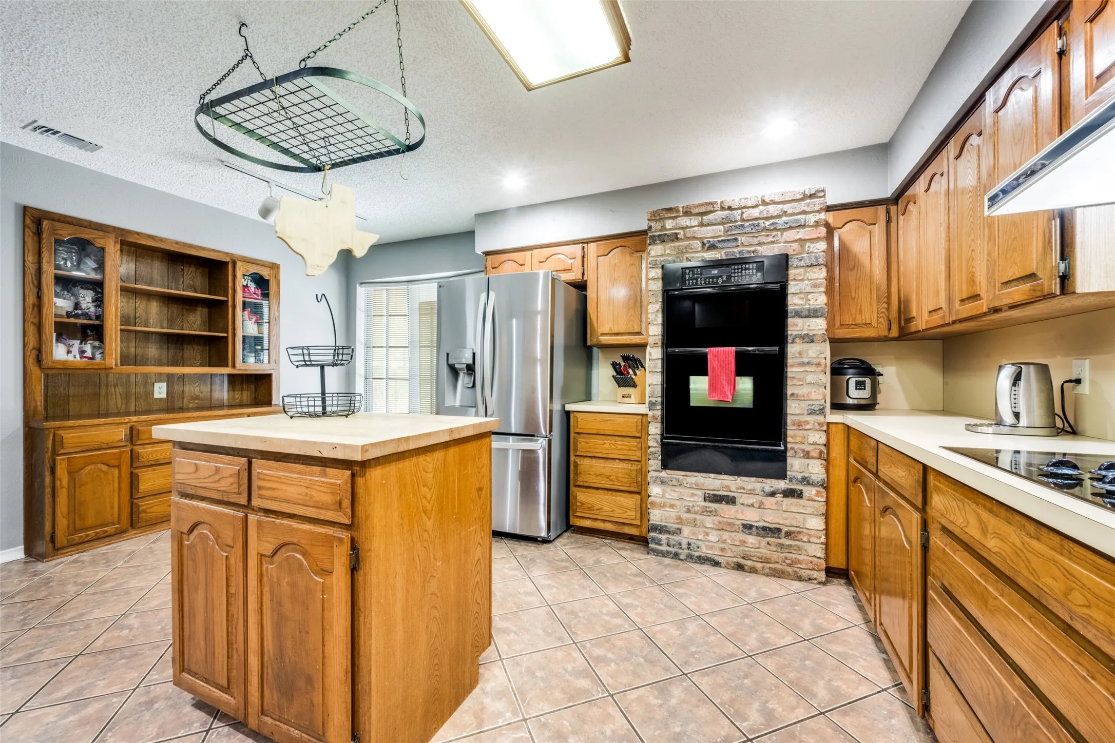 Kitchen featuring a built-in double oven within a brick surround, tile flooring, a central island with wood cabinetry, wood cabinetry throughout, and a stainless steel refrigerator