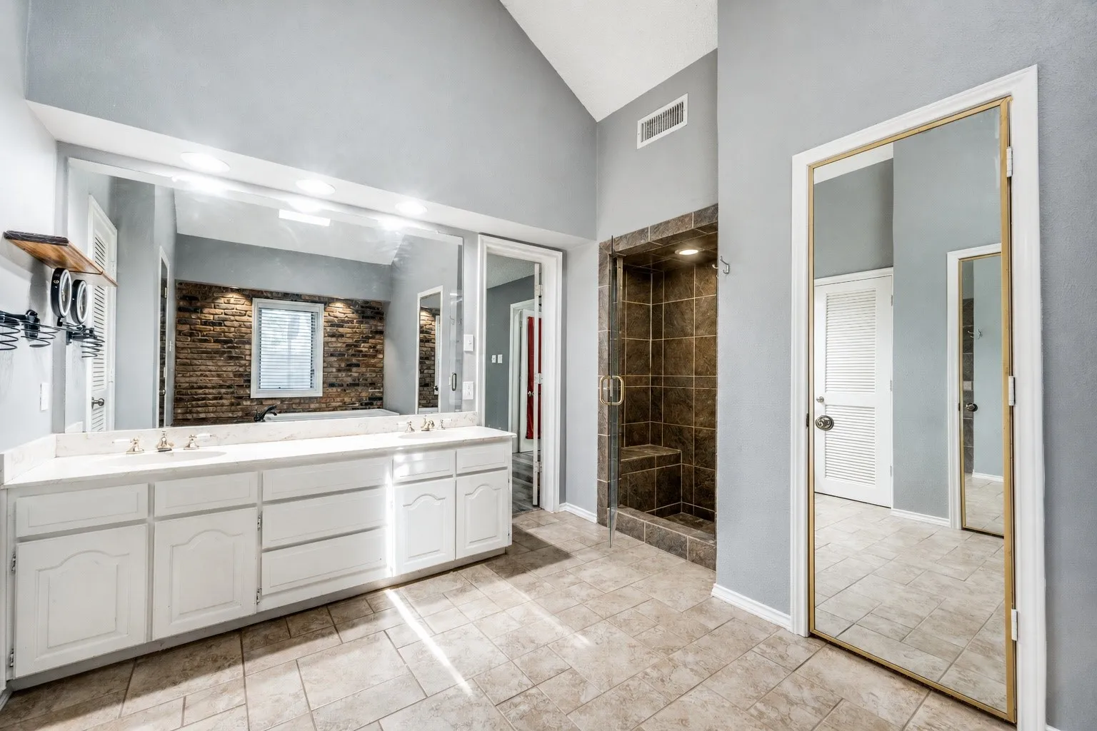 Spacious bathroom featuring a double vanity with white cabinetry and a light-toned countertop, a large frameless mirror with overhead lighting, a walk-in shower with brown tile surround and an integrated bench, and light-toned tile flooring
