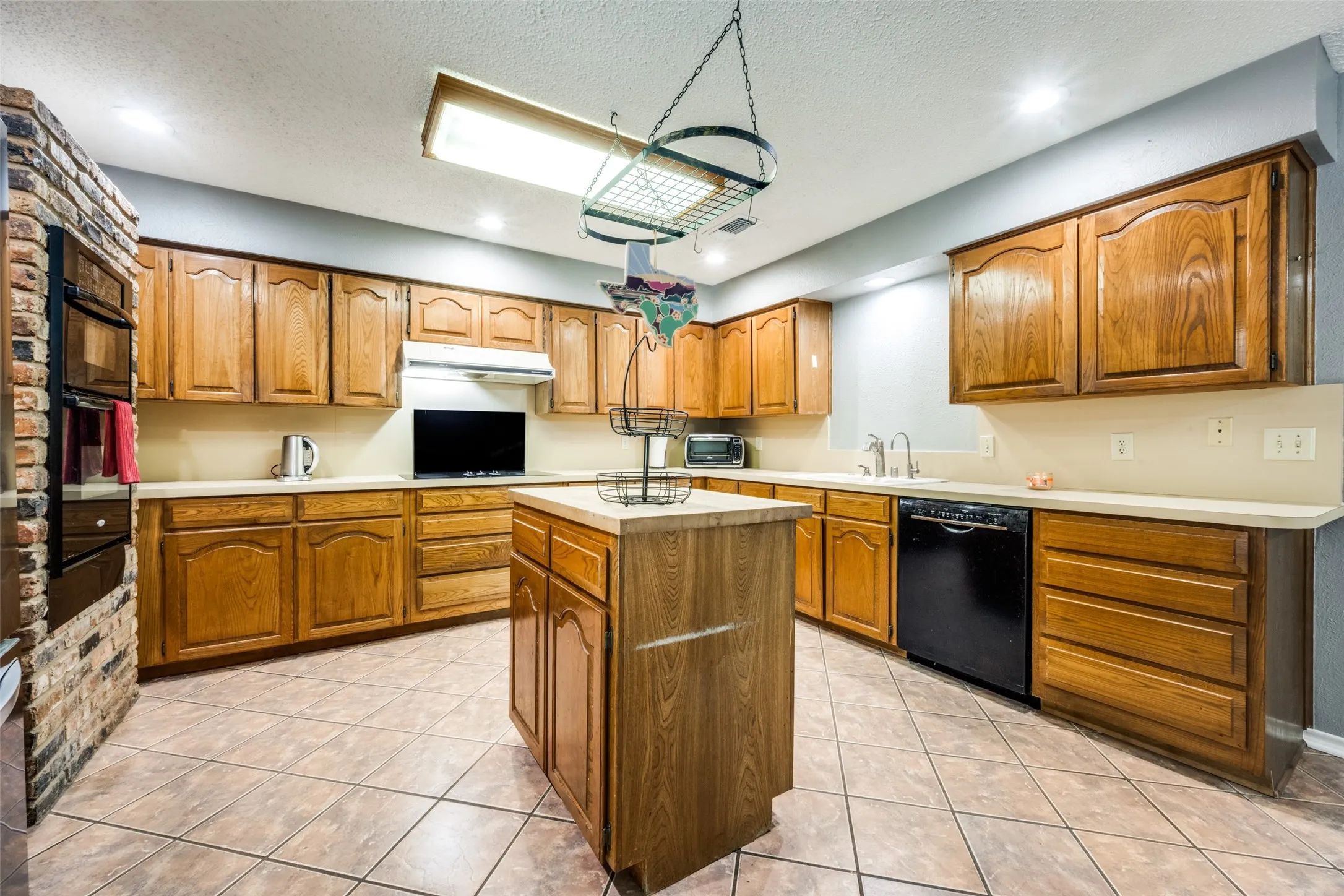 Kitchen featuring wood cabinetry, a central island, tile flooring, built-in ovens in a brick surround, and a black dishwasher