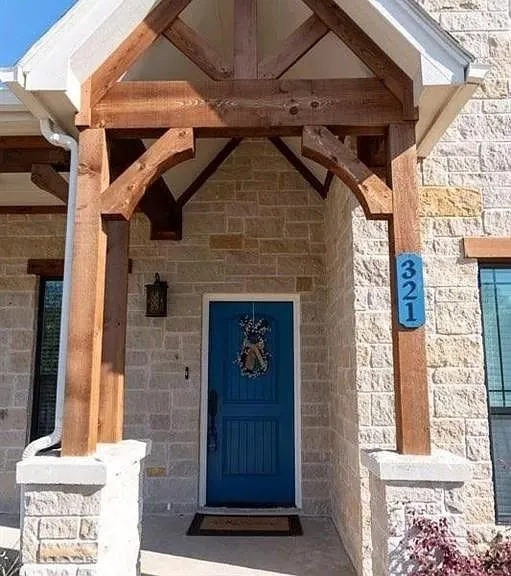 Entryway featuring a vibrant blue door, a stone facade, exposed wood structural elements, a built-in outdoor light fixture, and a concrete porch
