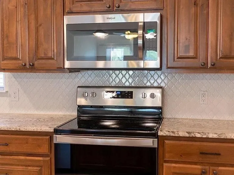 Kitchen featuring wood cabinetry, stainless steel microwave, stainless steel range with a black cooktop, white arabesque tile backsplash, and speckled countertops