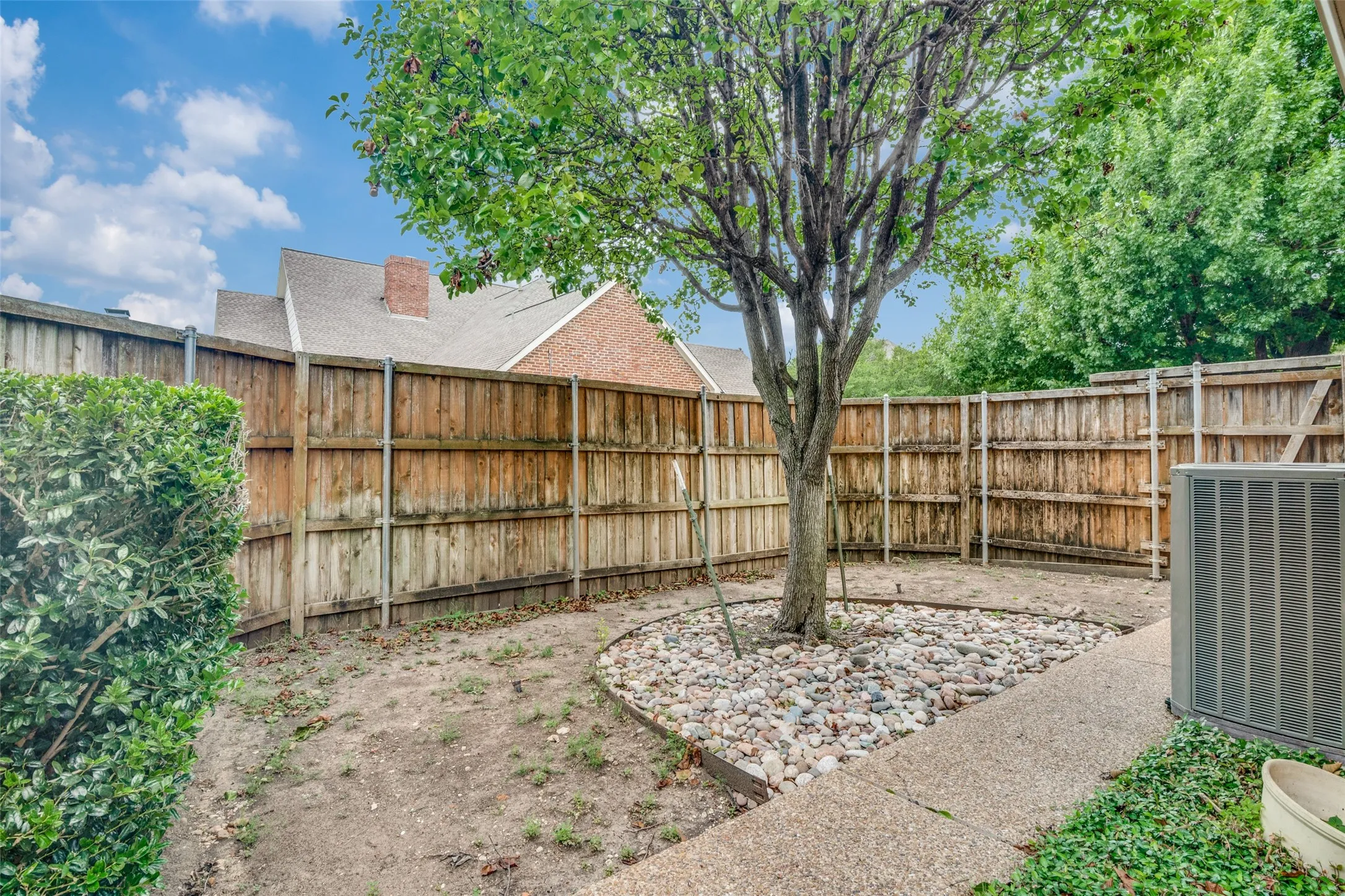 Private backyard featuring a mature tree with a rock garden base, concrete patio, wood privacy fencing, and established perimeter shrubbery
