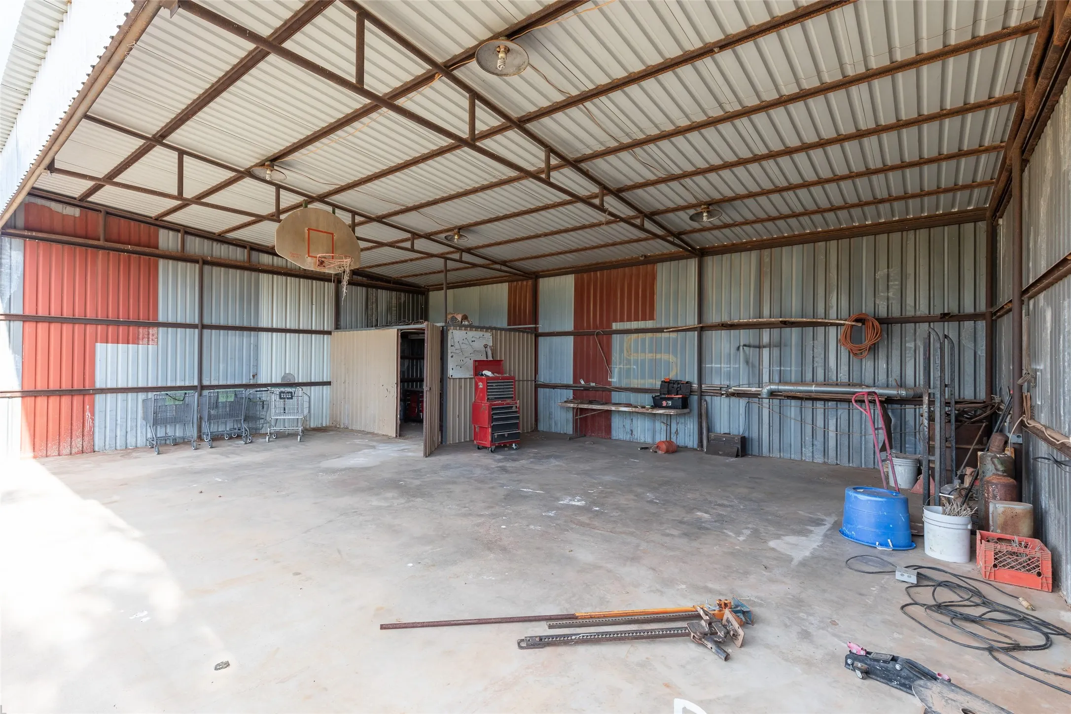 Open-sided metal structure featuring a corrugated roof, concrete flooring, and an integrated storage room with a wooden door