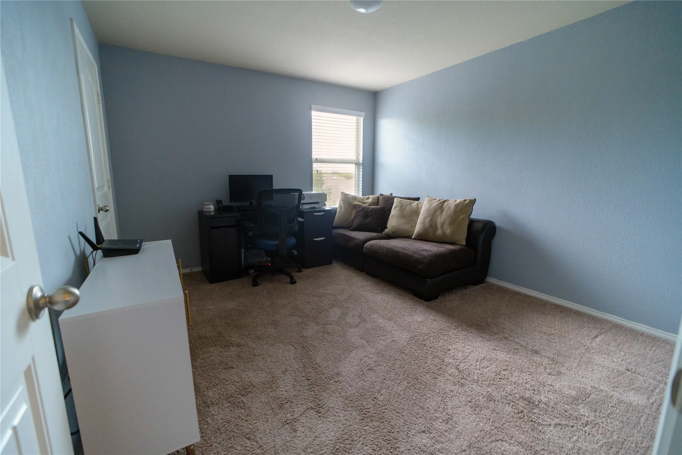 Carpeted interior room featuring light blue painted walls, a single window with horizontal blinds, and white baseboards. Used as an office.