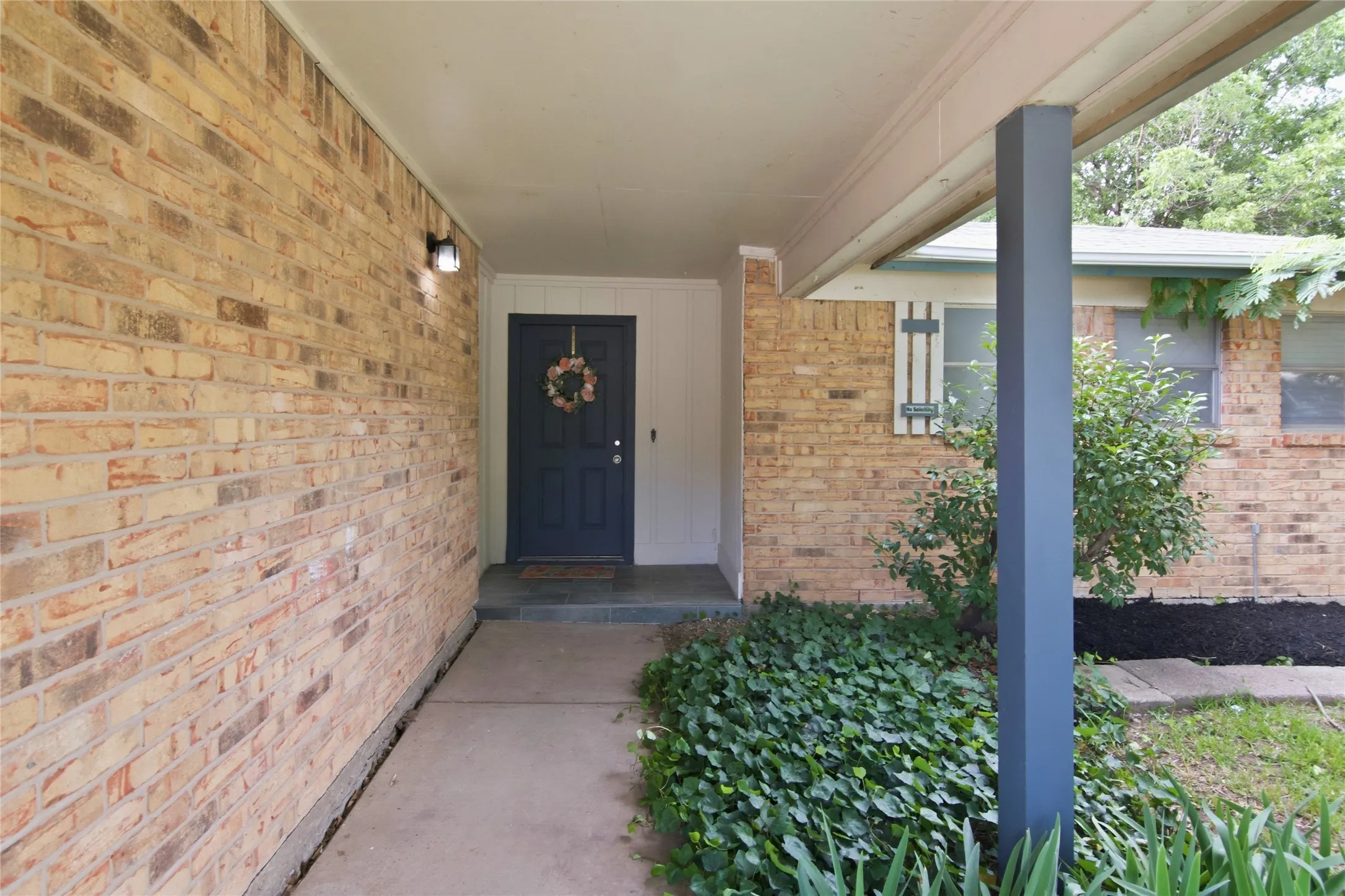 Covered entryway featuring a paneled front door, brick exterior, concrete walkway, and established landscaping