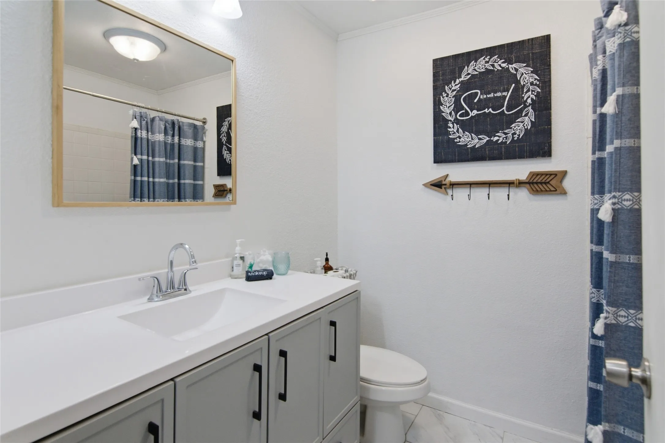 Bathroom featuring a vanity with a white countertop and gray cabinetry, a rectangular mirror with a wood-finish frame, a white toilet, and white tile flooring