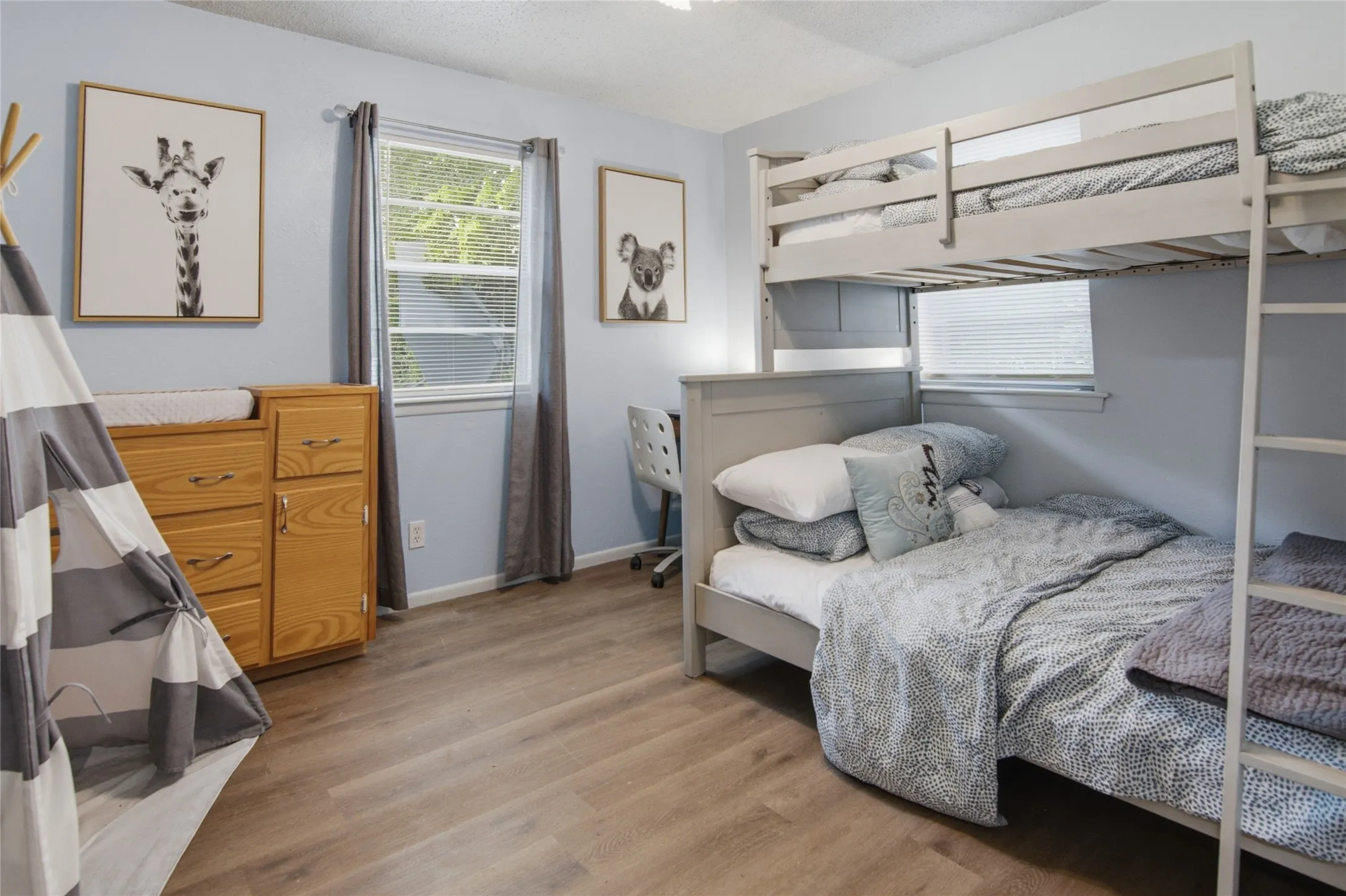 Bedroom featuring wood-finish flooring, two windows with blinds and curtains, and light blue wall paint