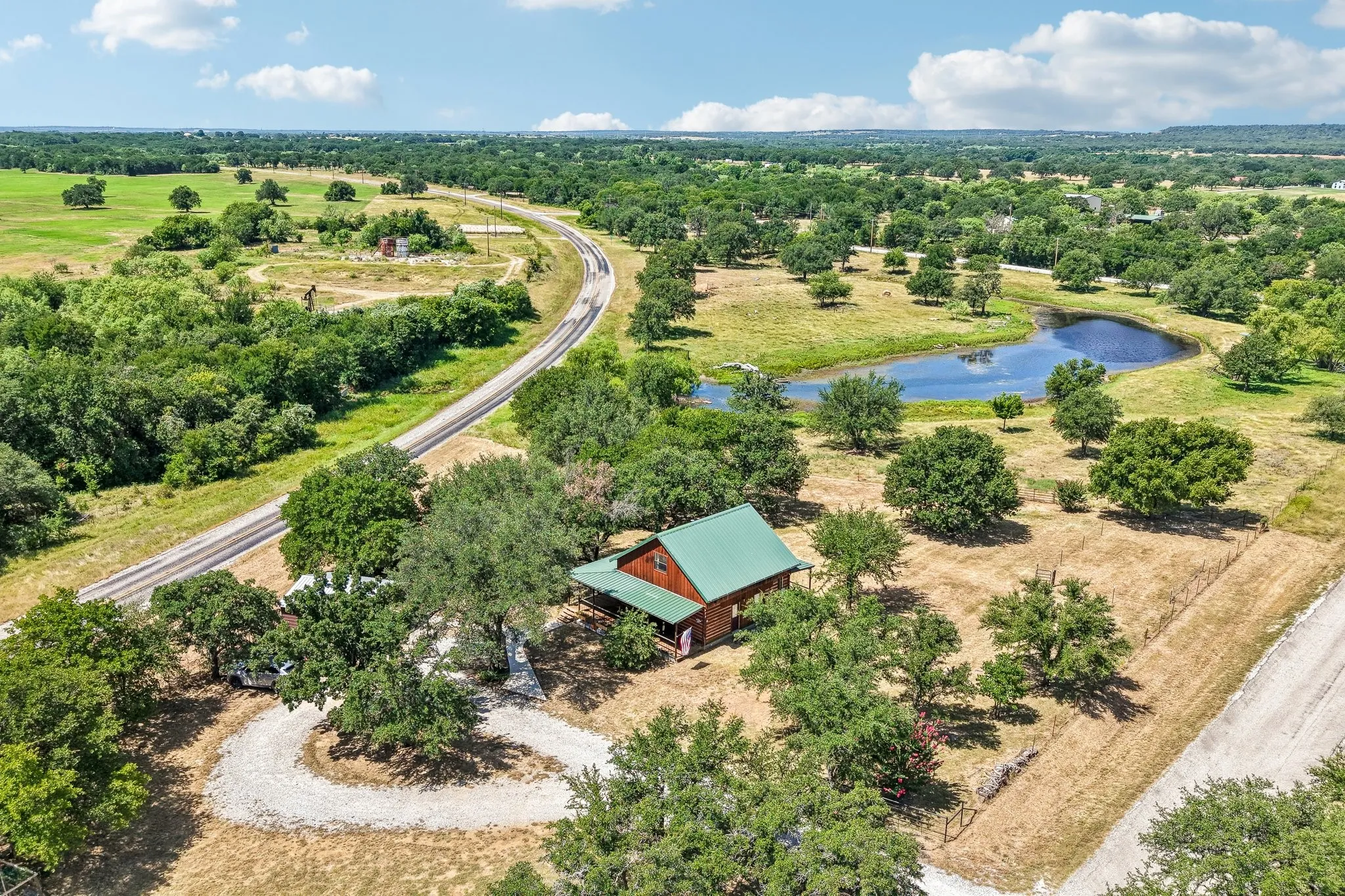 Log cabin with green metal roof, a curved gravel driveway, and a pond surrounded by extensive trees and open land