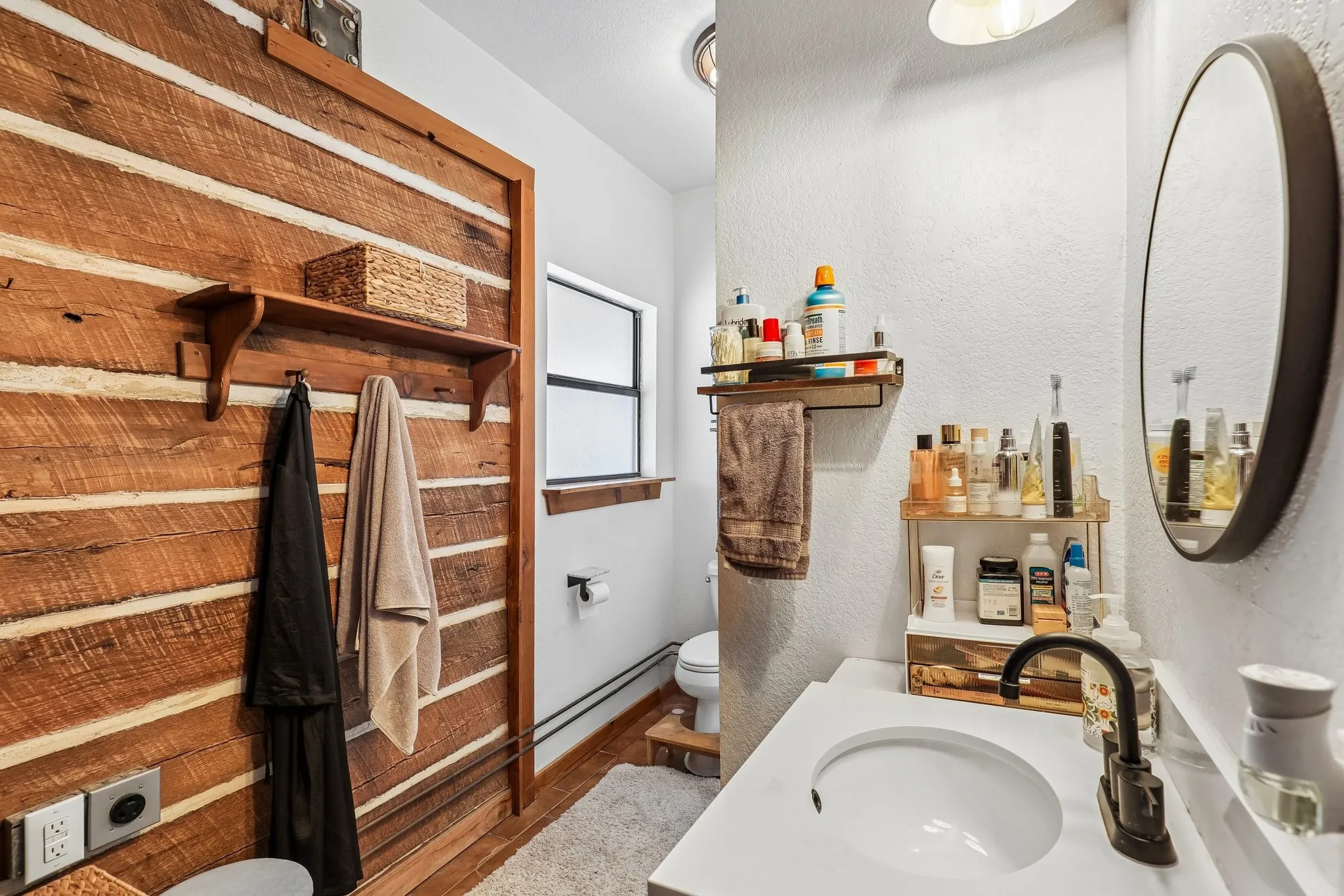 Bathroom featuring a wood plank accent wall, white vanity with integrated sink, black single-handle faucet, oval mirror, and a window with wood trim