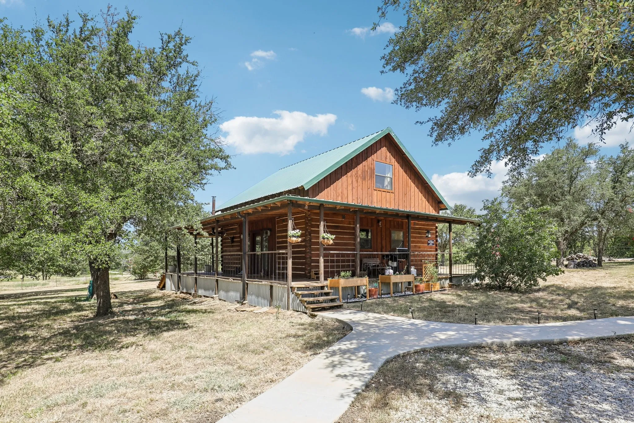 Log cabin exterior featuring a covered front porch, a green metal roof, natural wood siding, an upper-level window, and surrounding mature trees