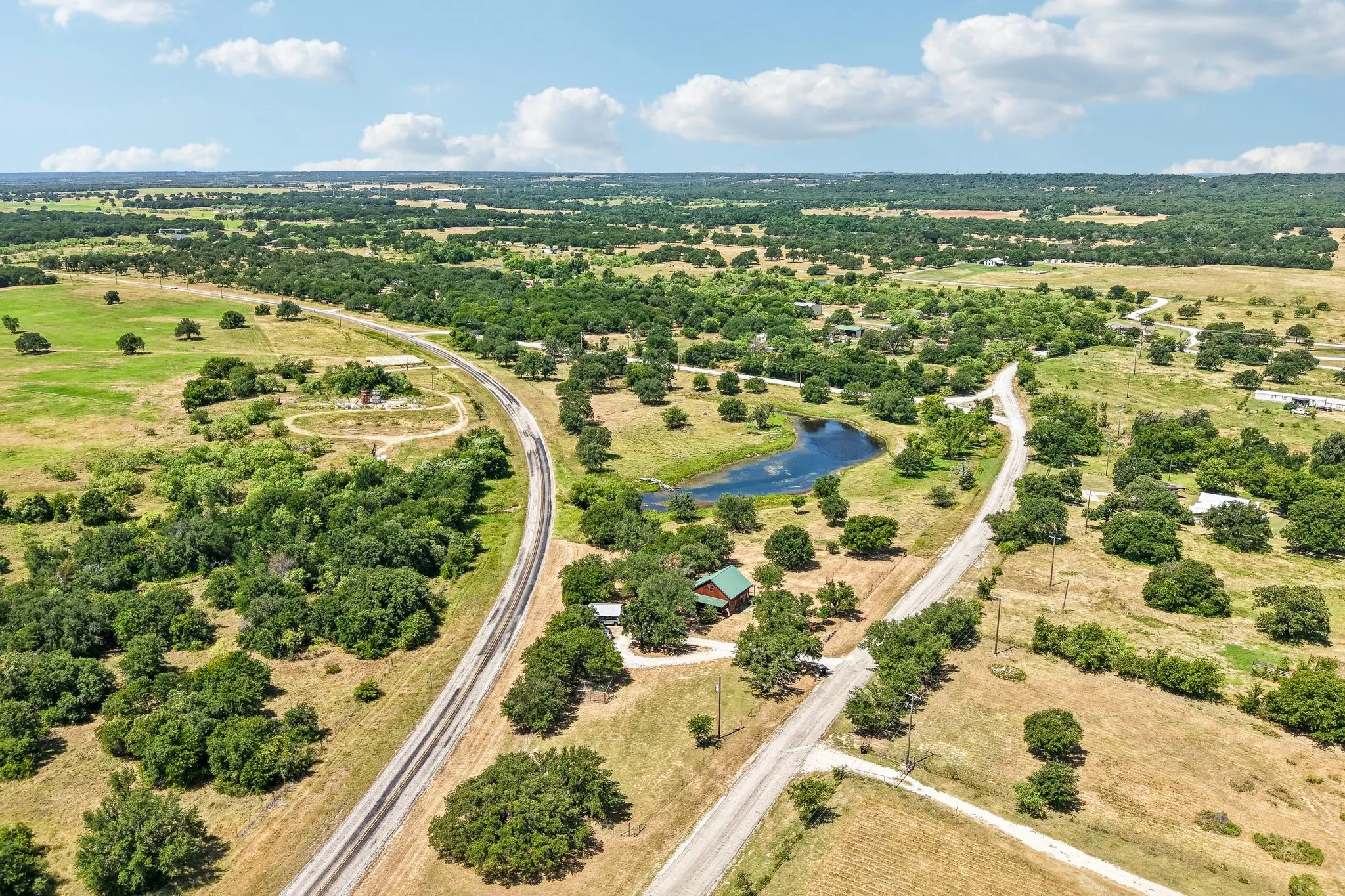 Expansive rural landscape featuring a winding paved road, numerous mature trees, a natural pond, a single-story structure with a green roof, and a gravel driveway